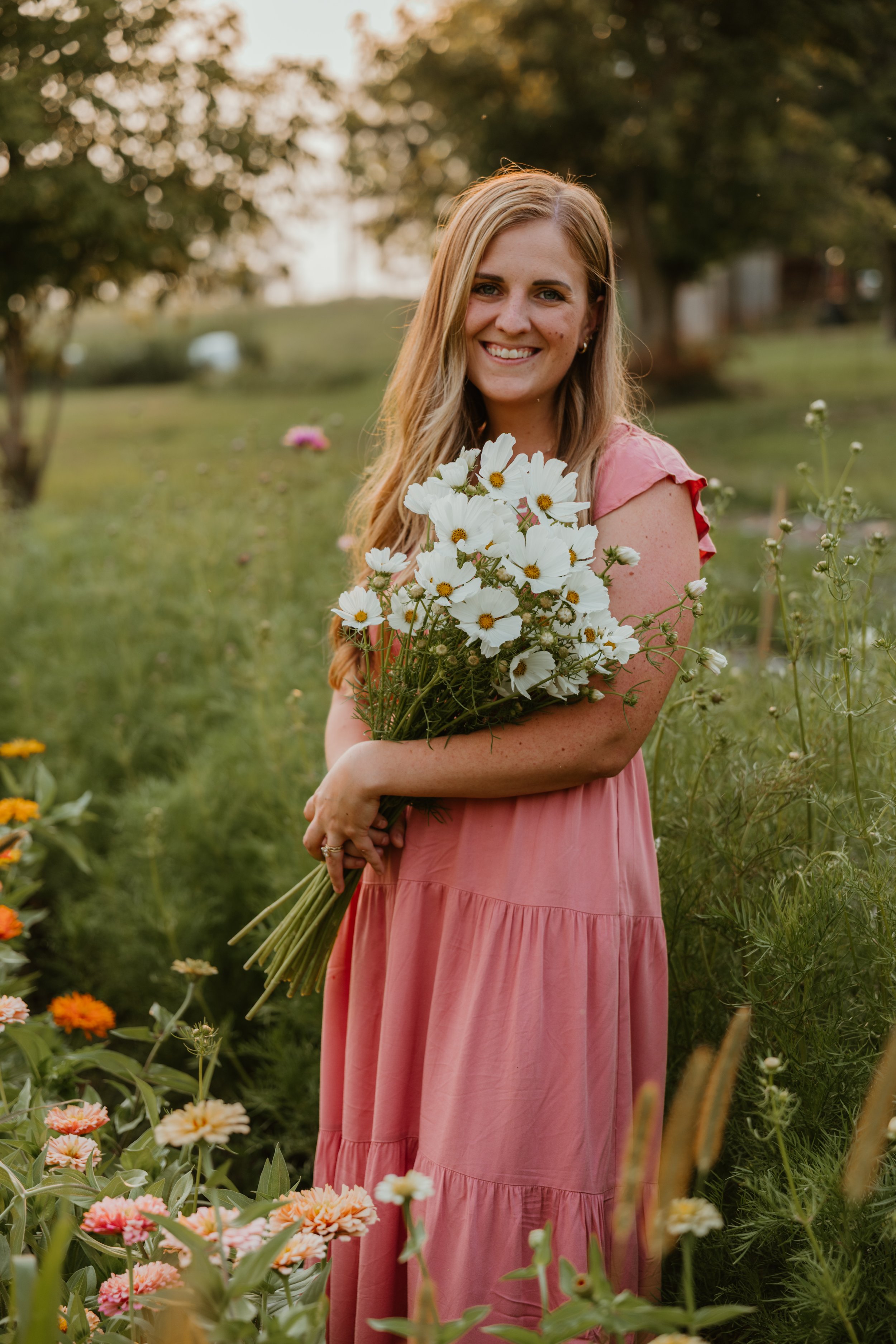 A smiling young woman in a pink dress standing in a garden, holding a bouquet of white cosmos, with colorful flowers and green foliage around her during sunset.