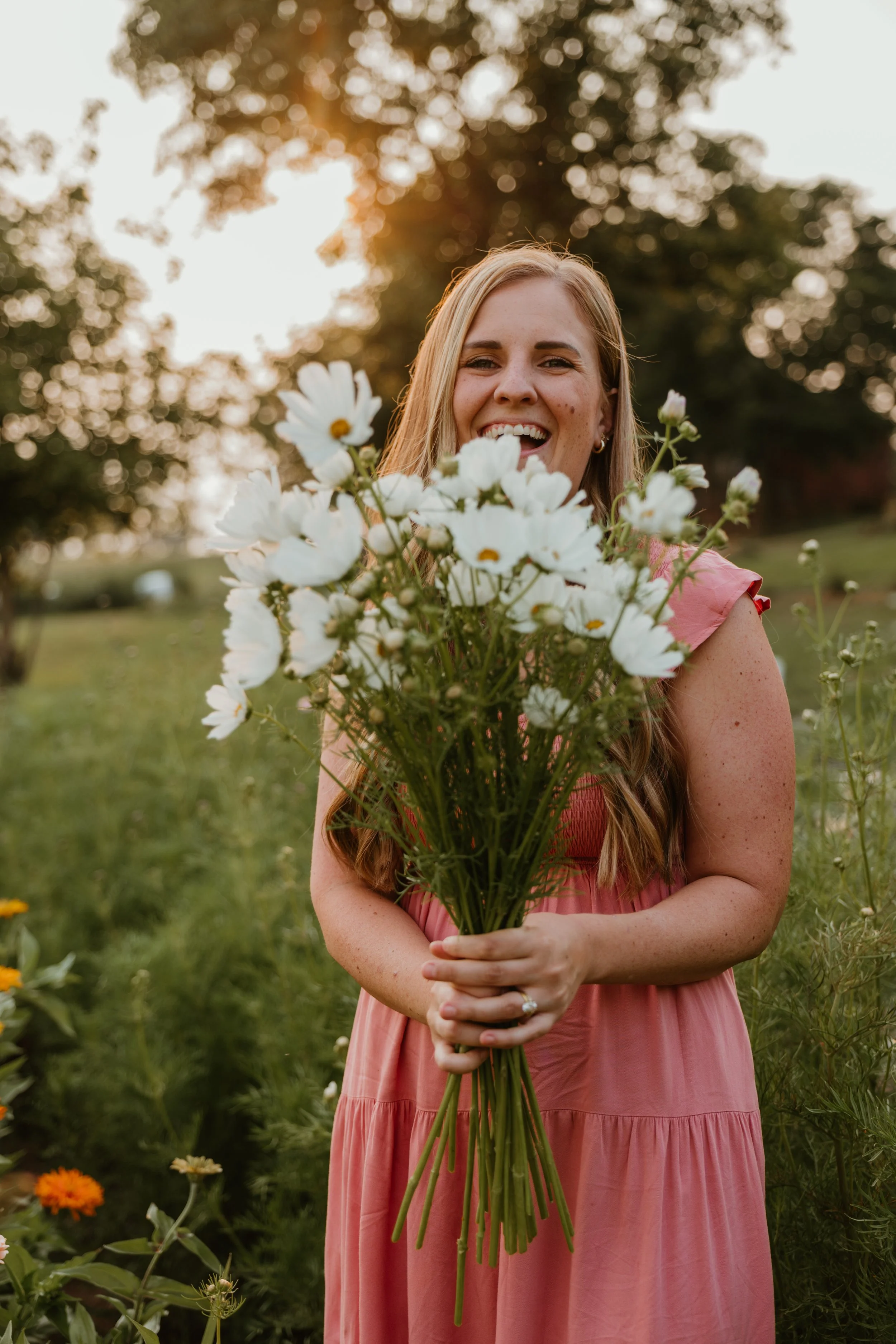 Woman in pink dress smiling and holding a bouquet of white cosmos in a garden at golden hour.