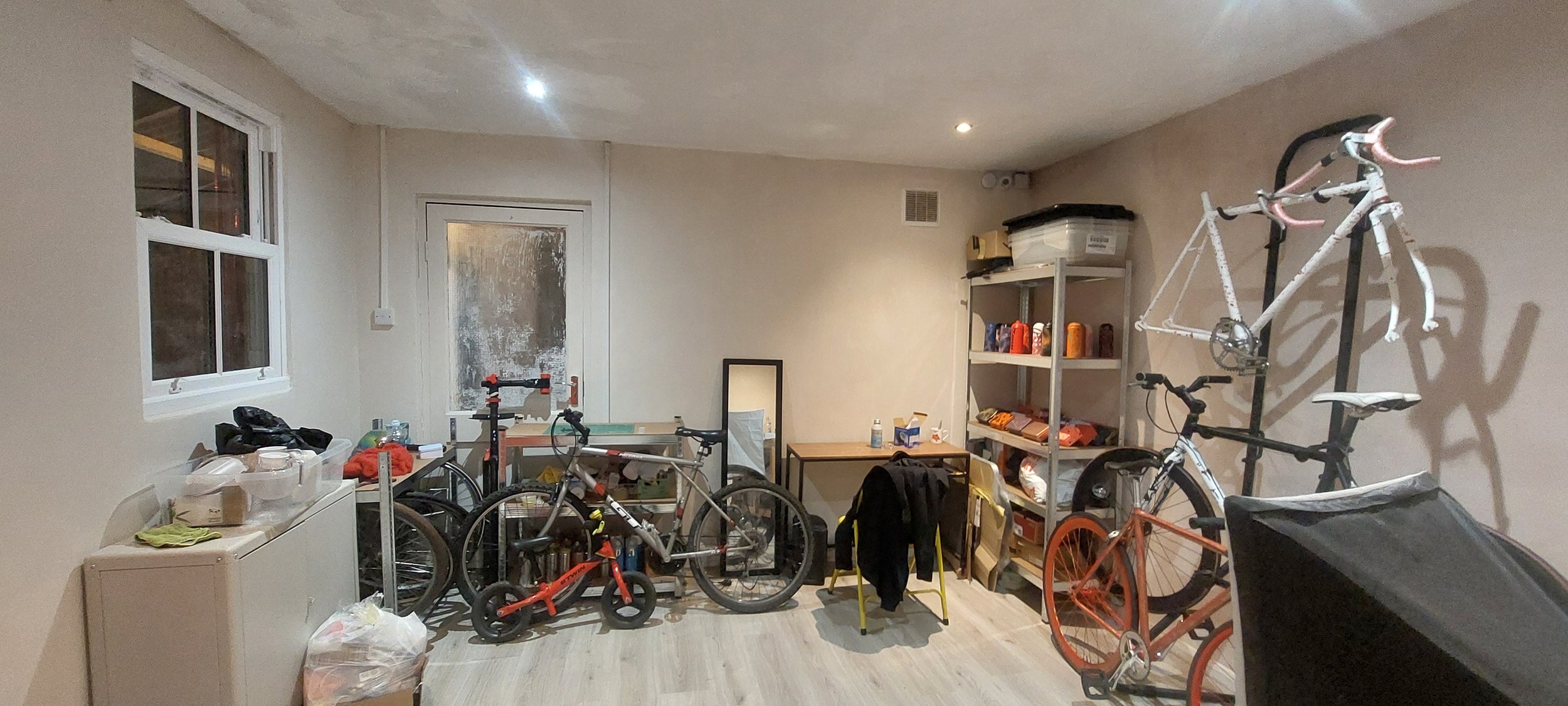 A cluttered room with bicycles, shelves, and various items on a table and floor.