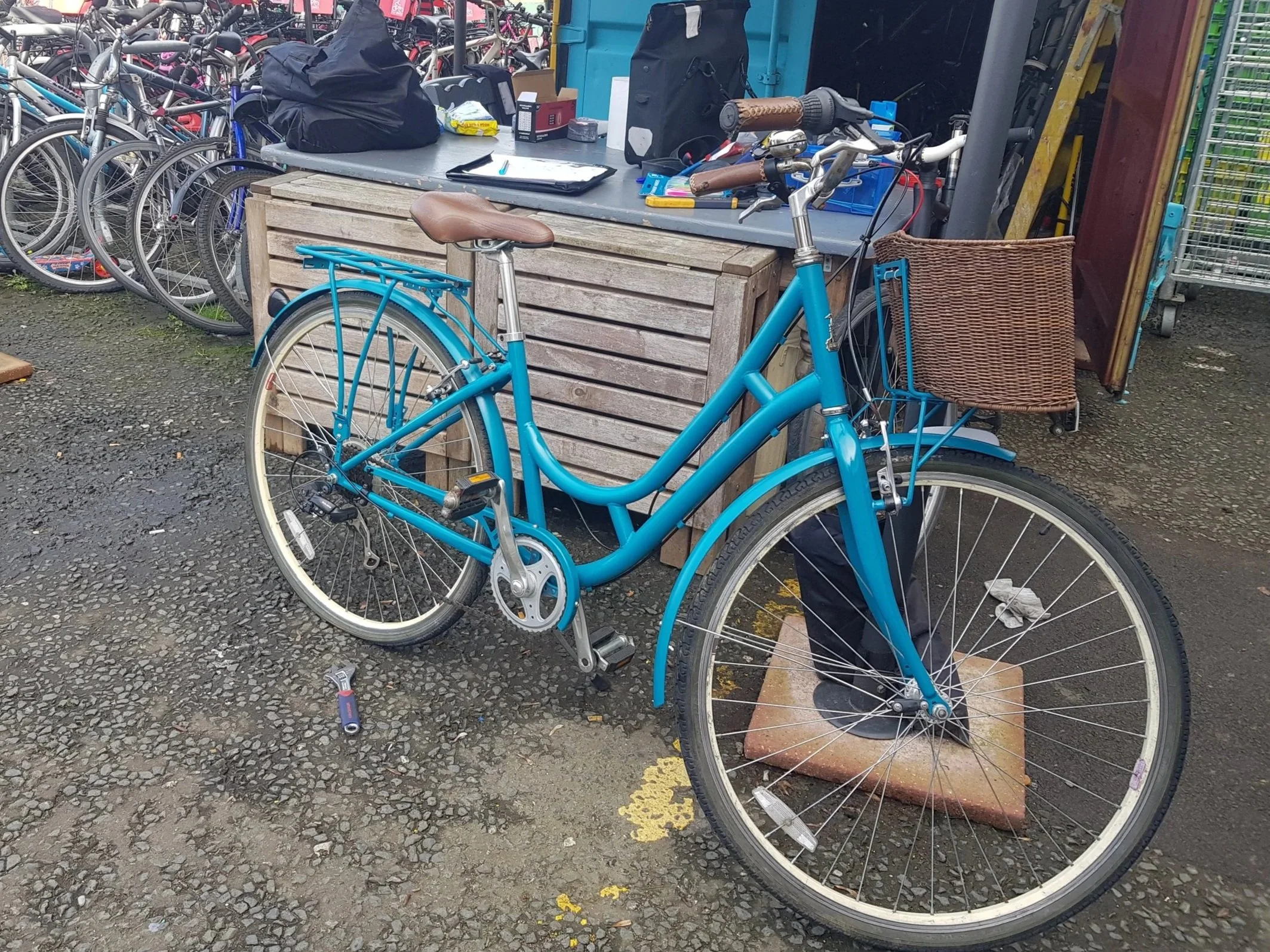 A blue bicycle with a brown leather seat and a wire basket parked on a brick at an outdoor bike shop, with other bikes and shop items in the background.