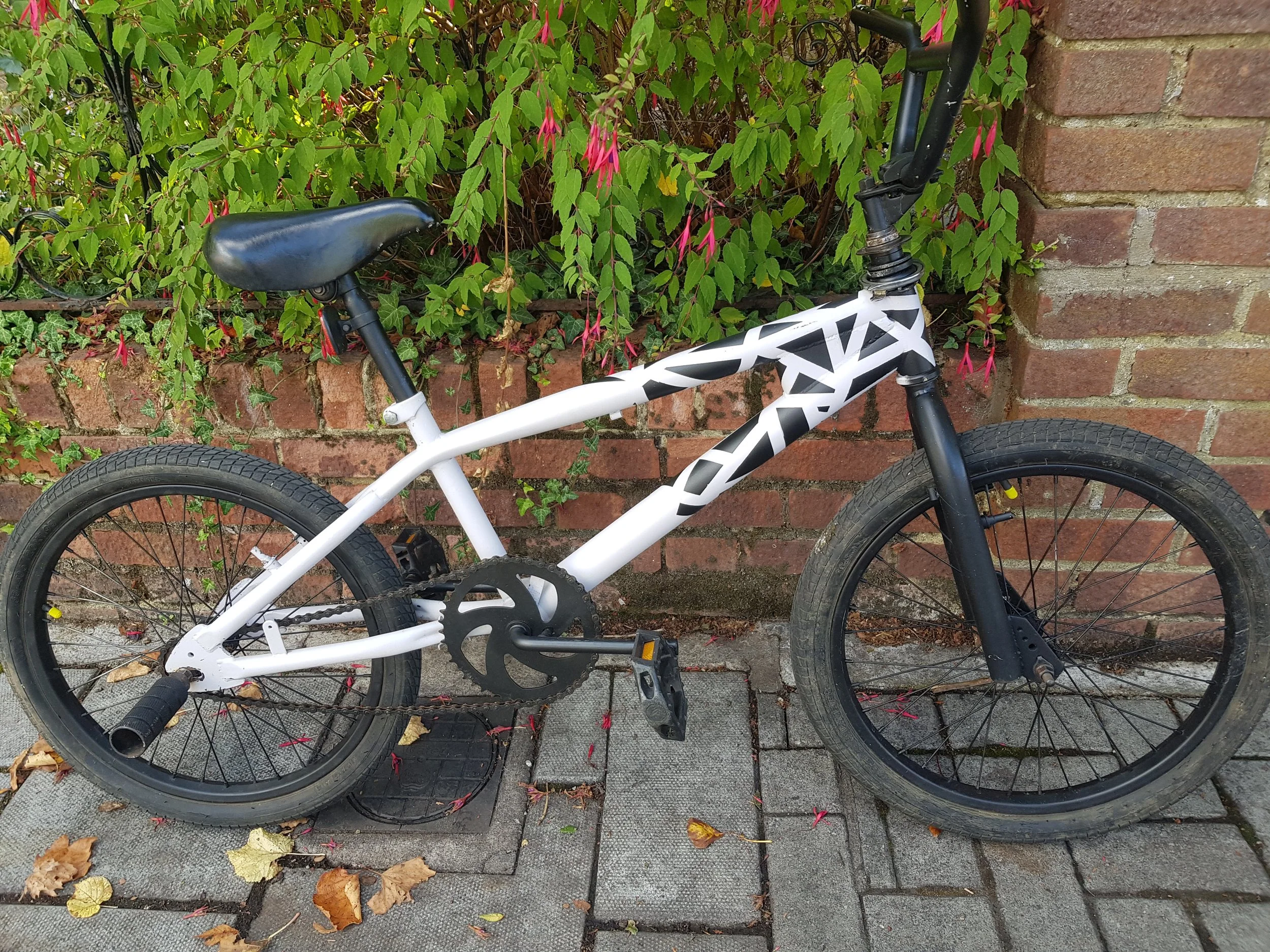 A black and white mountain bike with a black seat, black tires, and a black front fork, parked against a brick wall with green foliage and pink flowers above it, on a sidewalk with fallen leaves around.
