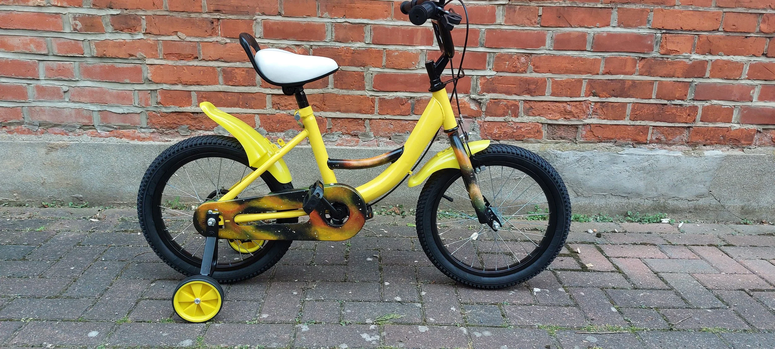 Yellow children's bicycle with training wheels and a black and orange spray paint design, parked on a brick sidewalk against a red brick wall.