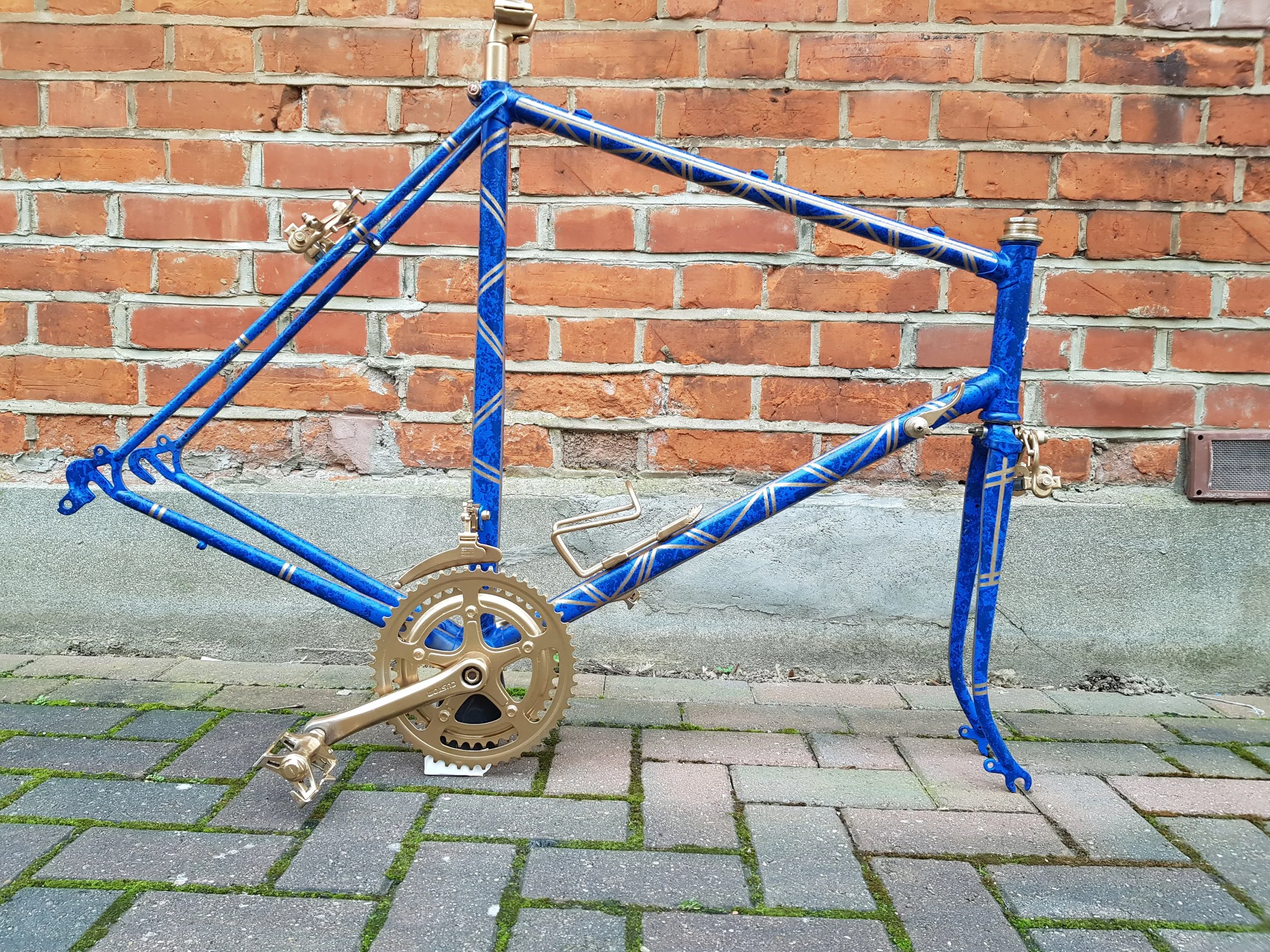 Blue bicycle frame with gold components, placed upright on a sidewalk against a brick wall.