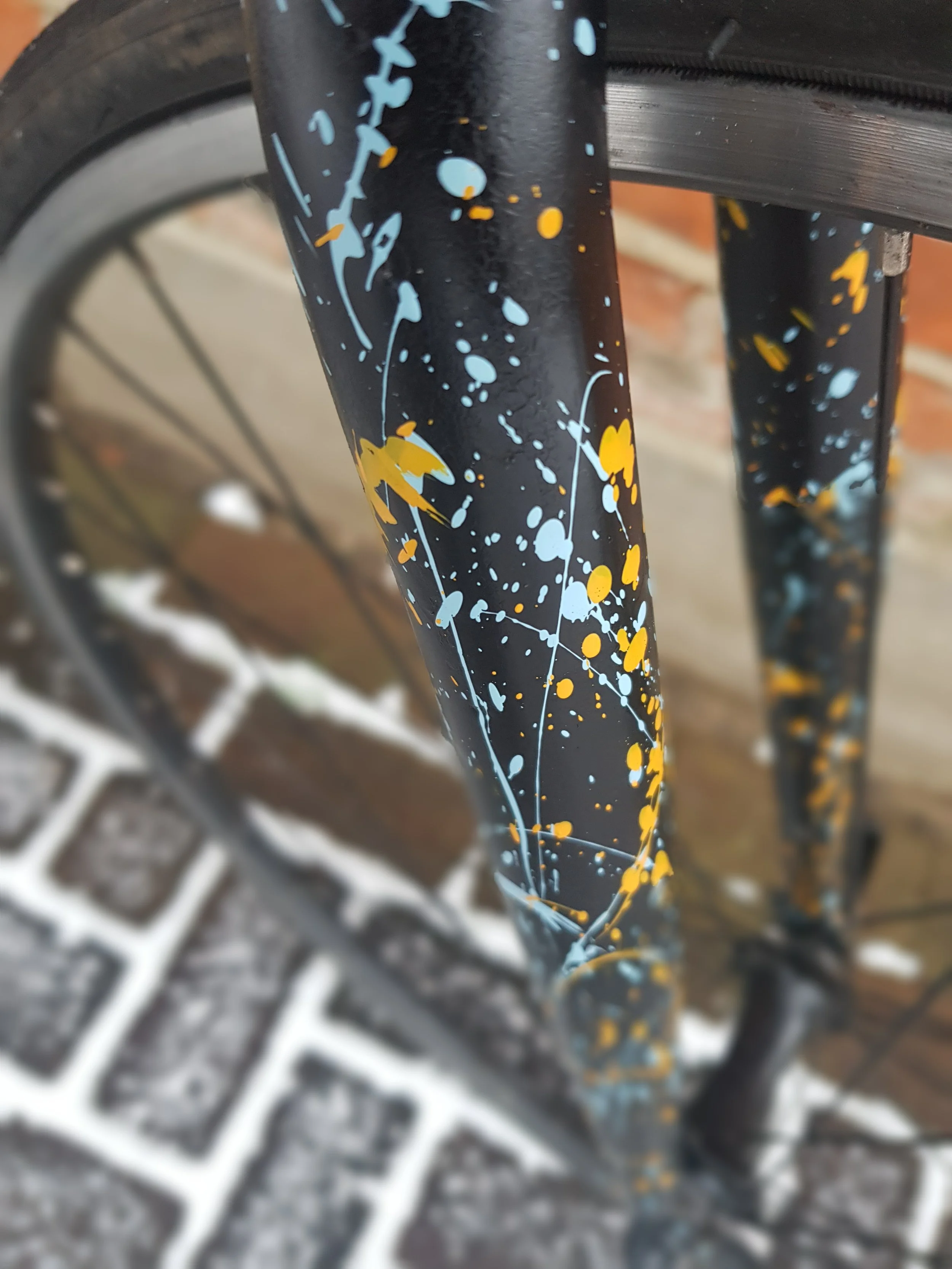 Close-up of a bike fork with a splatter paint design in blue and yellow on a black background, with a brick and concrete surface in the background.