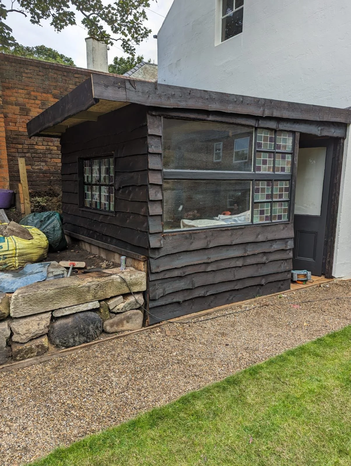 Small structure with black wooden siding and a large window, attached to a house, with construction materials and tools nearby.