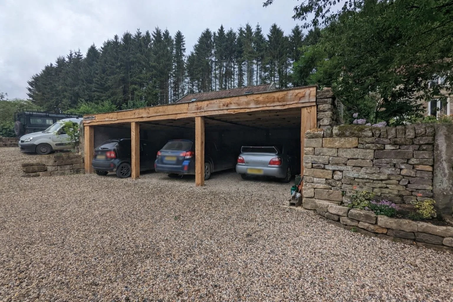 A stone and wood carport with three parked cars, gravel driveway, and trees in the background.