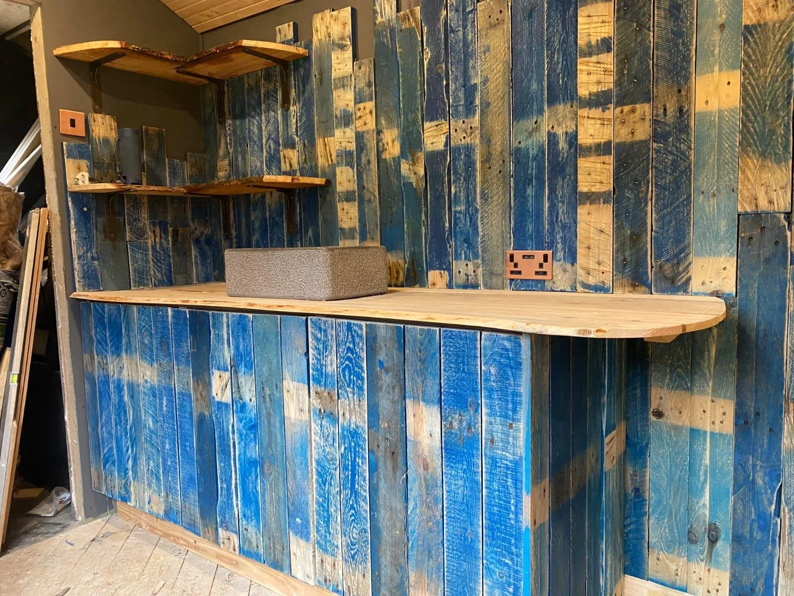 A rustic kitchen or bar area featuring a wooden counter with a blue and natural wood-paneled wall backdrop. There are three corner shelves mounted on the wall, and a stone container sitting on the counter. Two electrical outlets are visible on the wall, and the setting appears to be in a workshop or unfinished space.