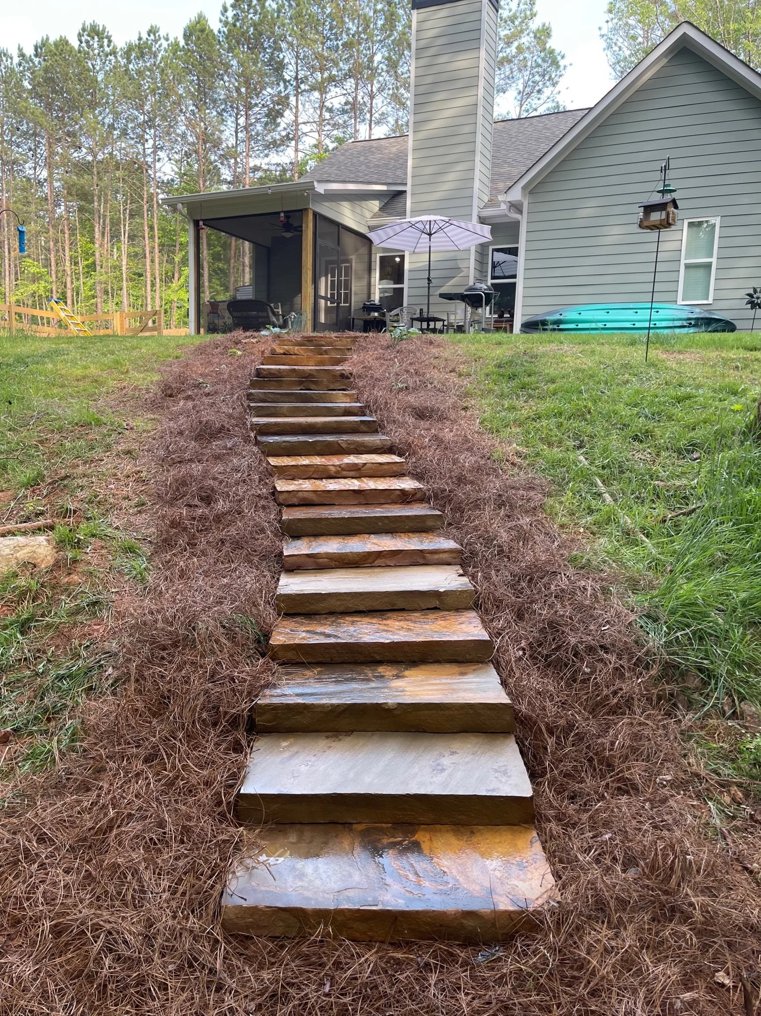 Wooden steps leading up a grassy hill to a backyard patio with furniture, a grill, and a screened porch, with trees in the background.