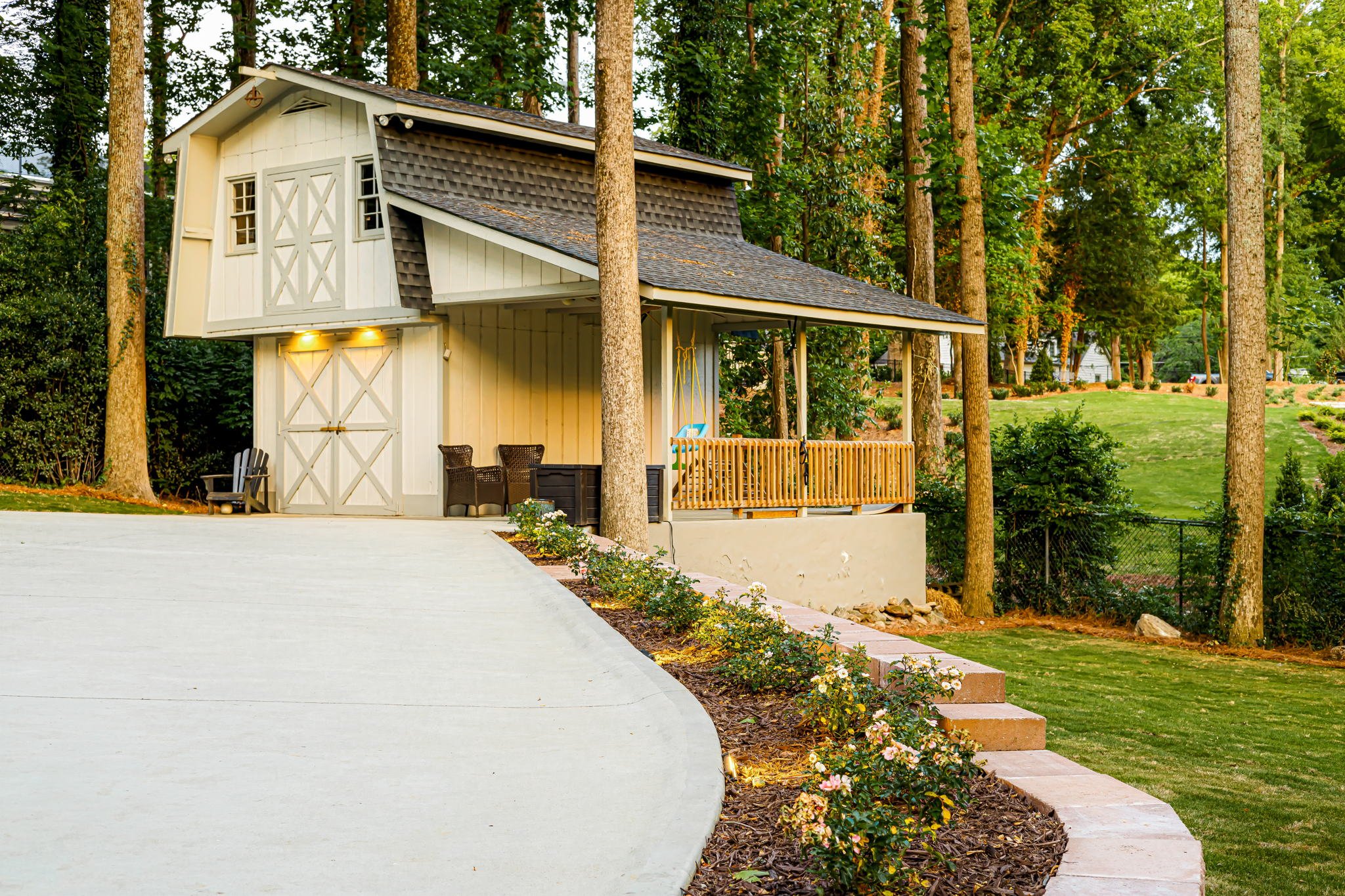 A small white and beige shed with a covered porch and black wicker chairs, surrounded by tall trees and a well-maintained garden and lawn.