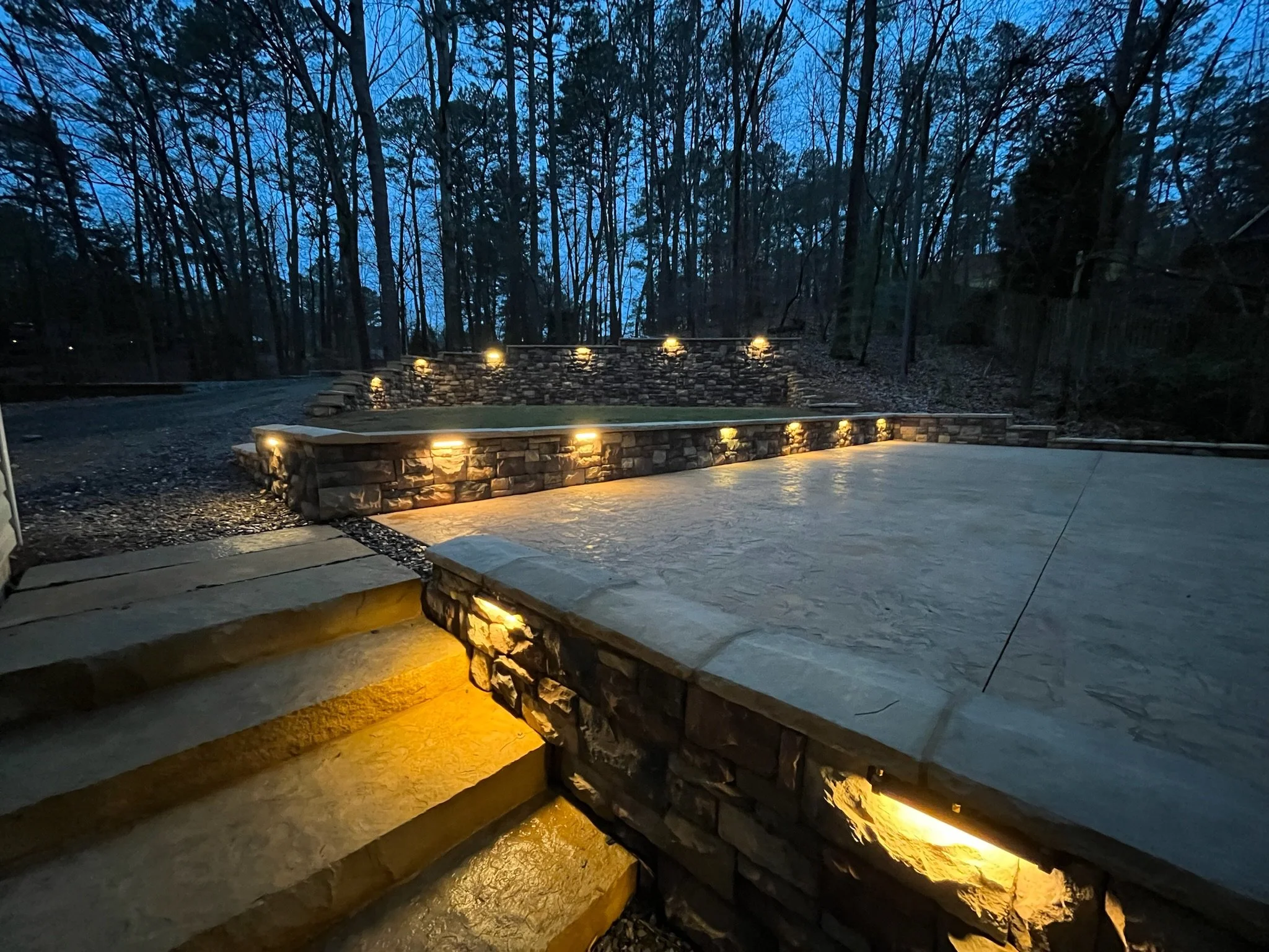 Outdoor backyard with stone steps and a flat surface, illuminated by small ground lights, surrounded by a wooded area with tall, leafless trees, during dusk or early evening.
