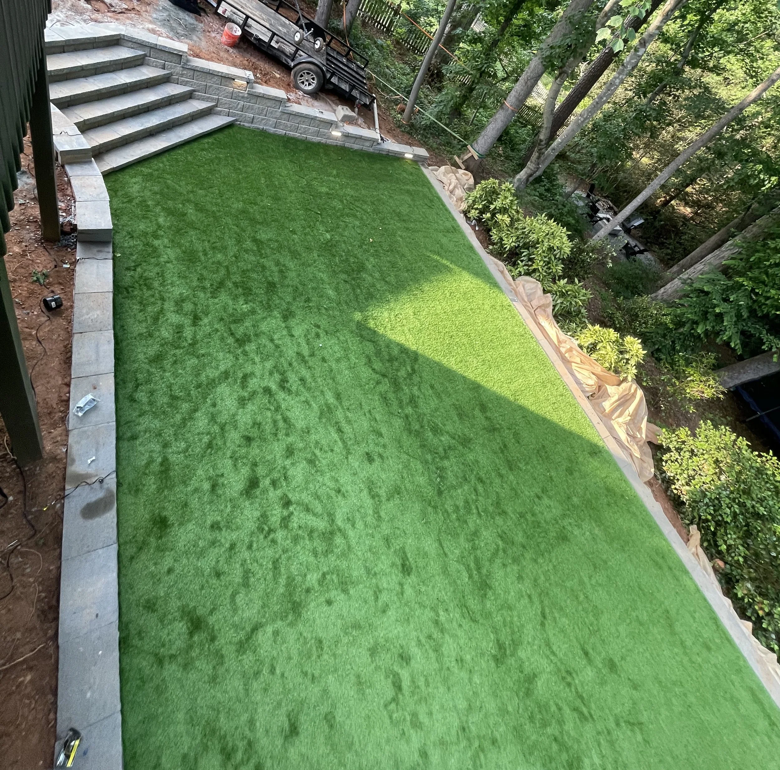 A backyard with newly installed bright green artificial grass, concrete steps on the left, and a landscaped area with trees and shrubs on the right.