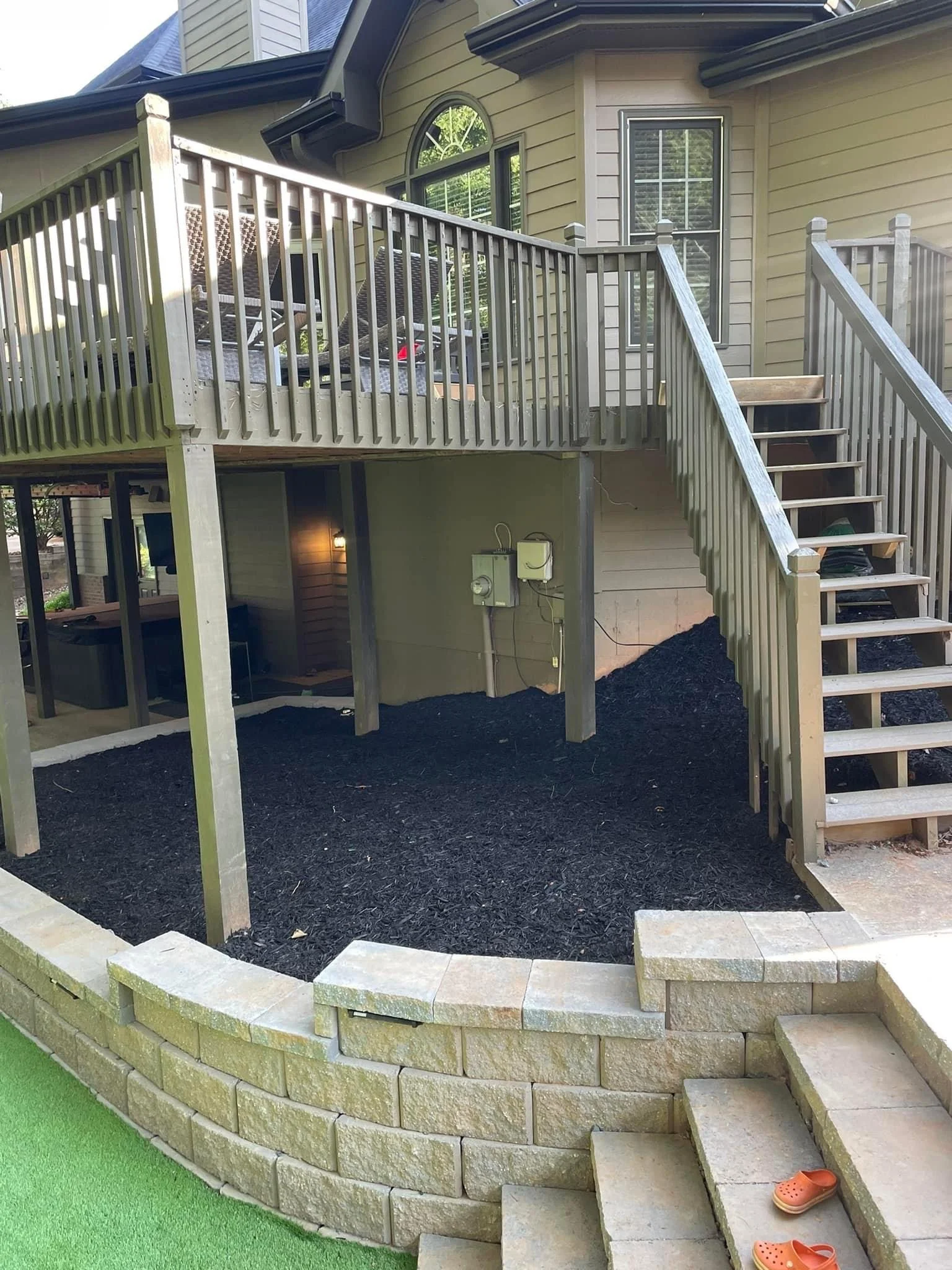 Backyard view showing a two-story house with a wooden deck and stairs, a black mulch bed, a stone retaining wall, and a small section of artificial grass. Crocs are on the steps.