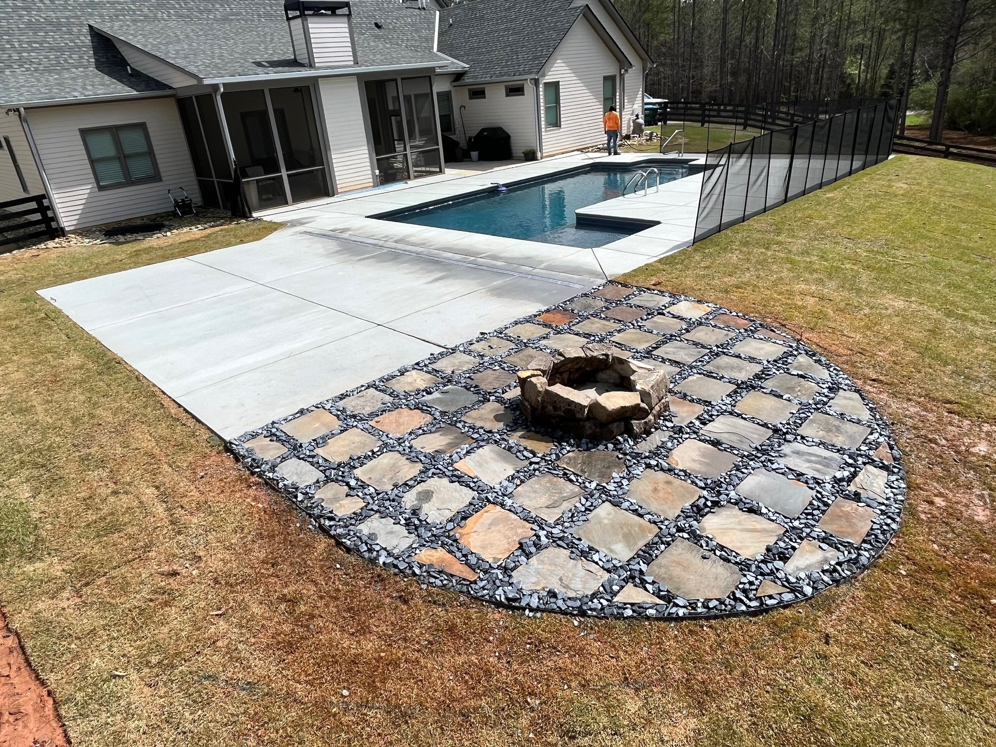 A backyard with a swimming pool, a fire pit area with stone and gravel, and a concrete patio adjacent to a house with an enclosed porch.