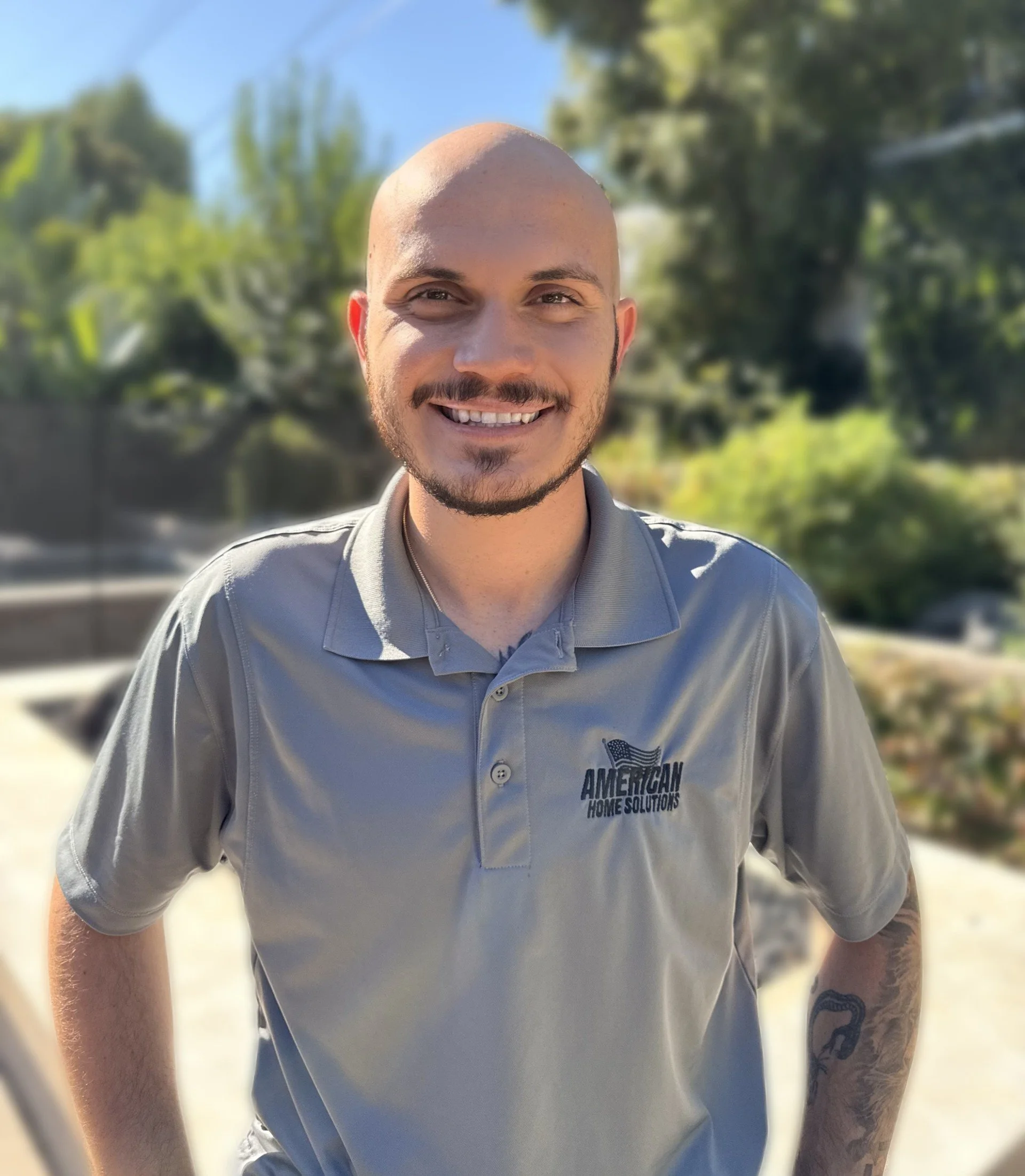 Man smiling outdoors wearing a gray polo shirt with "American Home Solutions" logo, with trees and a sunny sky in the background.