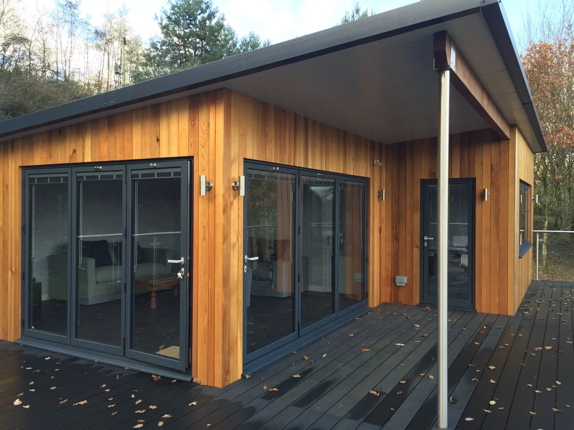 A modern wooden house with large glass sliding doors and a covered terrace, surrounded by trees.