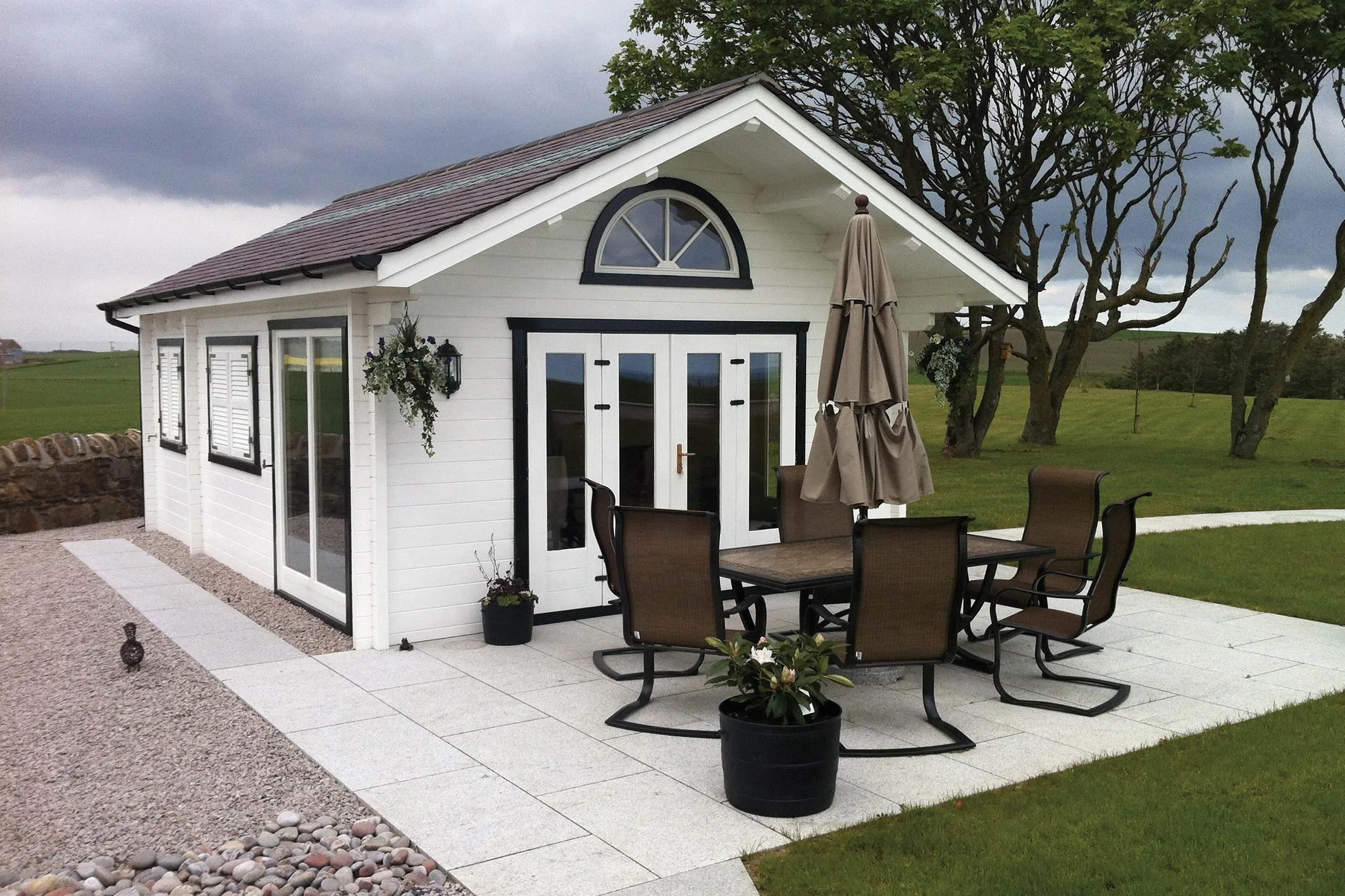 A small white shed with black trim, double glass doors, and an arched window above. It is surrounded by a paved patio with outdoor furniture including a table with a folded umbrella, and potted plants. There are trees and grassy fields in the background under a cloudy sky.