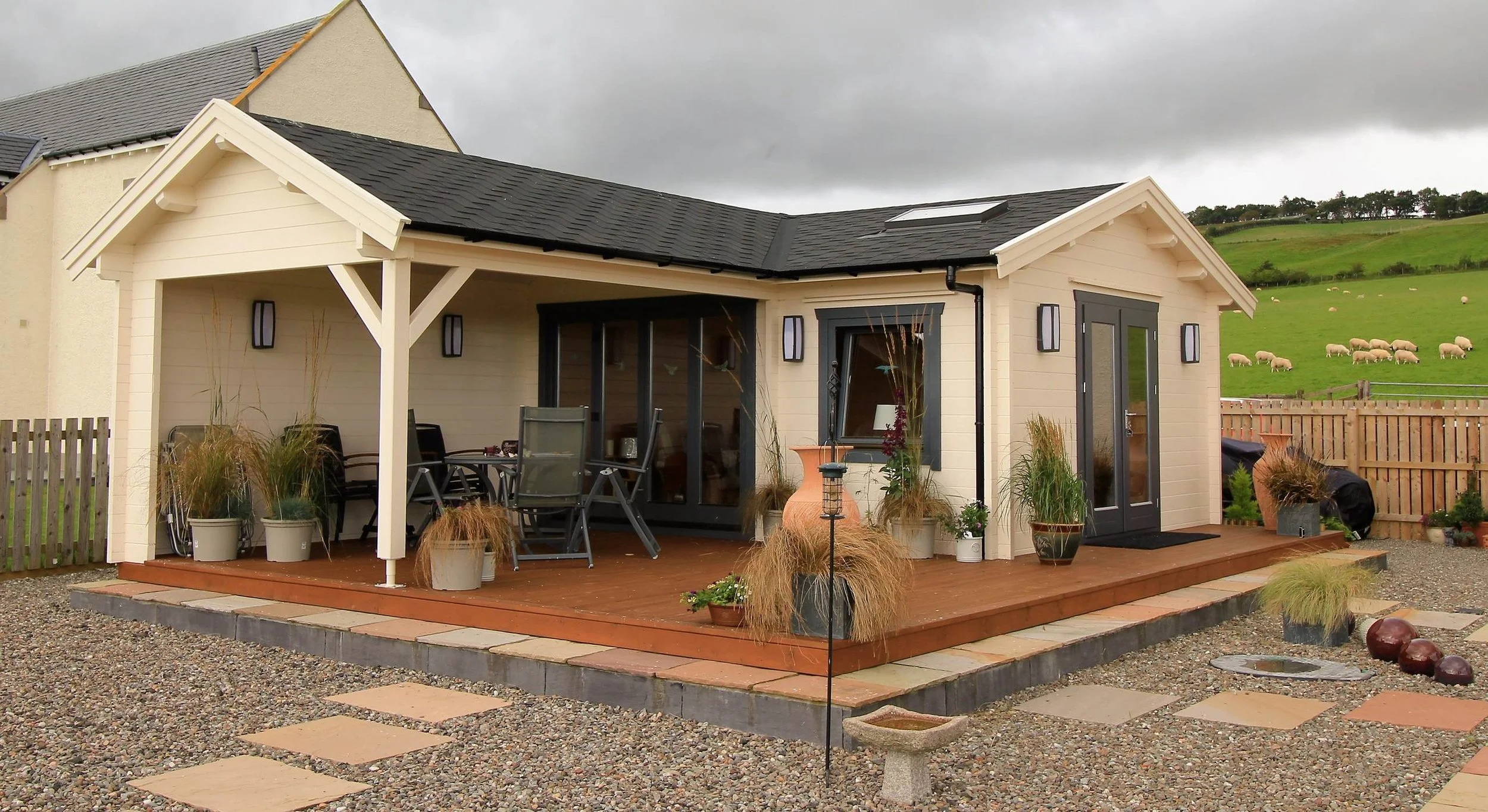 A cozy backyard patio with potted plants, outdoor furniture, and a wooden deck, with a grassy hillside and sheep grazing in the background.