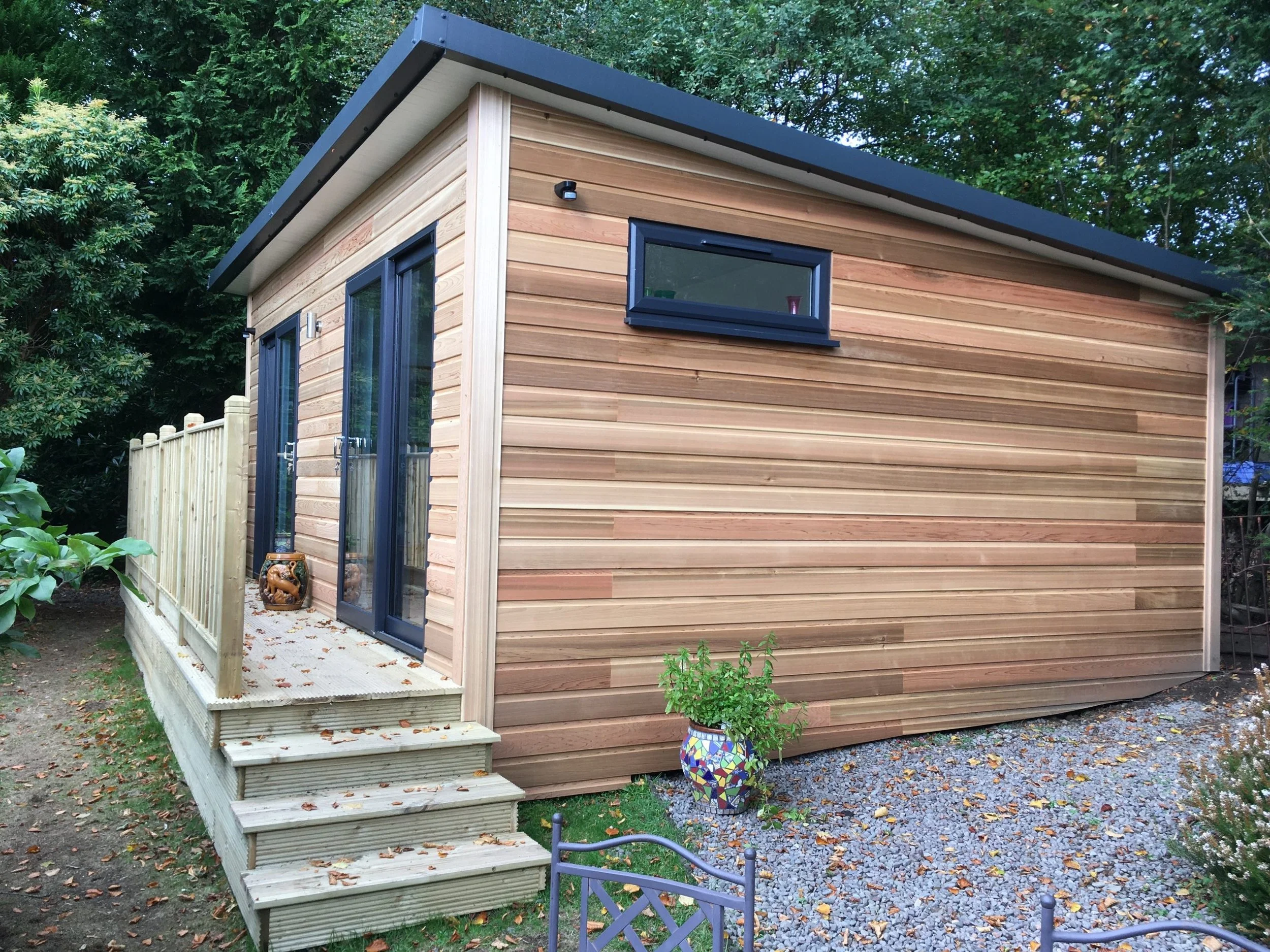 A small wooden house with a dark gray sloped roof, a sliding glass door on the front, a small horizontal window on the side, surrounded by green trees, with a wooden deck and steps, and a decorative plant pot outside.