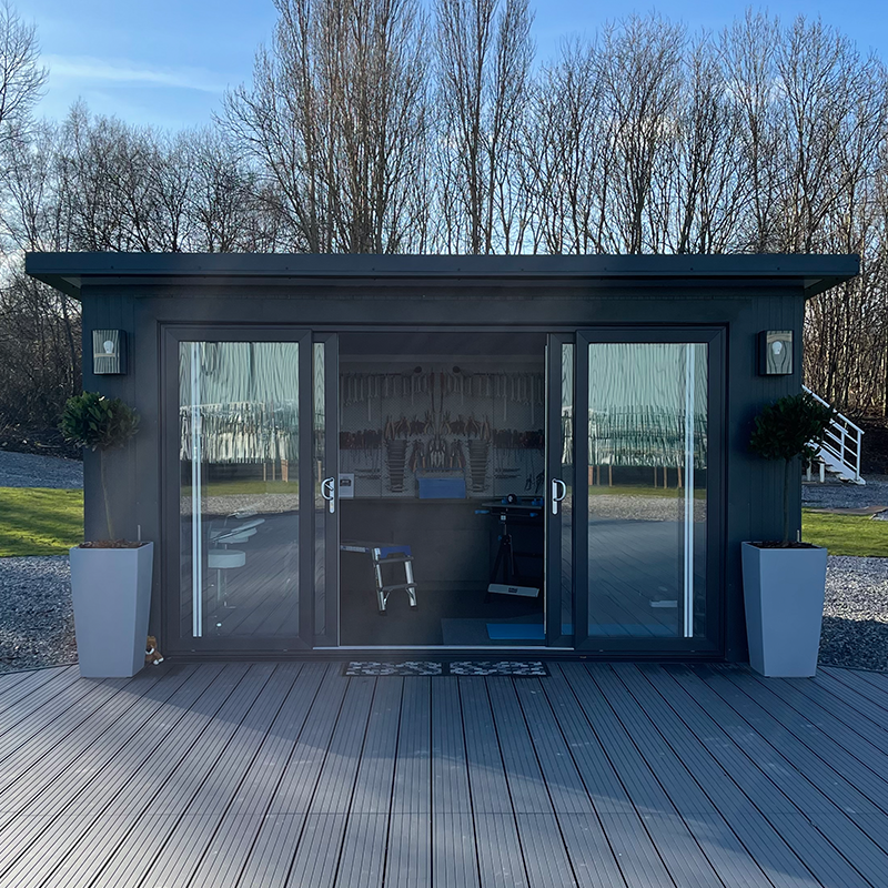 A small black shed with sliding glass doors, potted plants on each side, and outdoor lighting, located on a wooden deck in a backyard with trees.