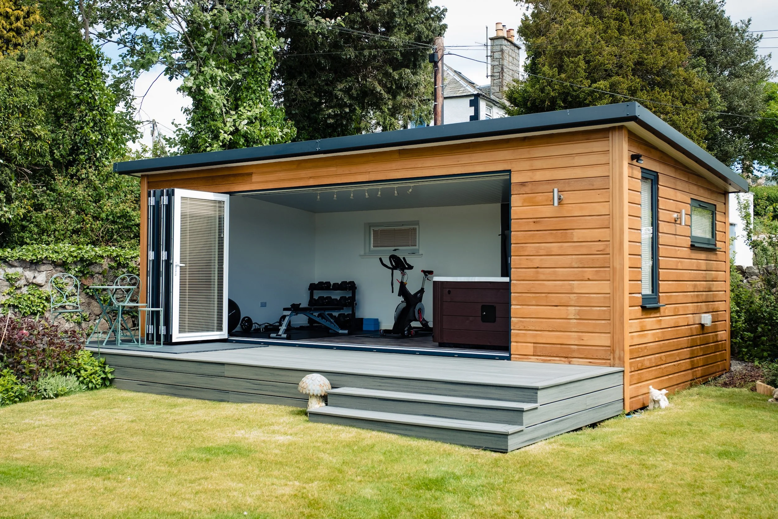 A small, modern backyard shed with open folding doors revealing a fitness area with exercise equipment. The shed is made of wood with a gray trim, set on a wooden deck with steps leading down to a green lawn. Surrounding greenery includes bushes and trees.