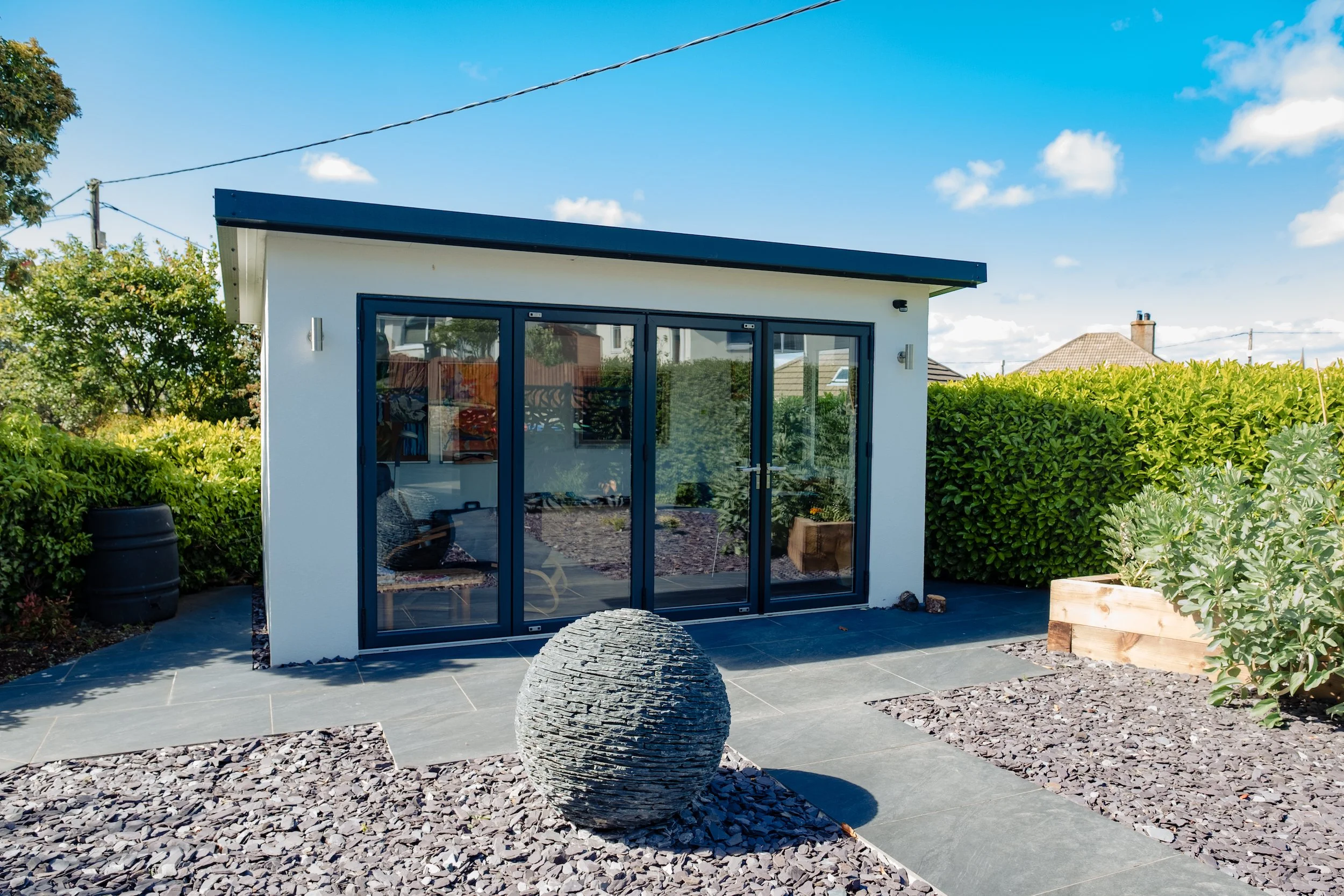 Small modern shed with glass doors in a backyard, surrounded by green bushes and a gravel garden, under a blue sky.
