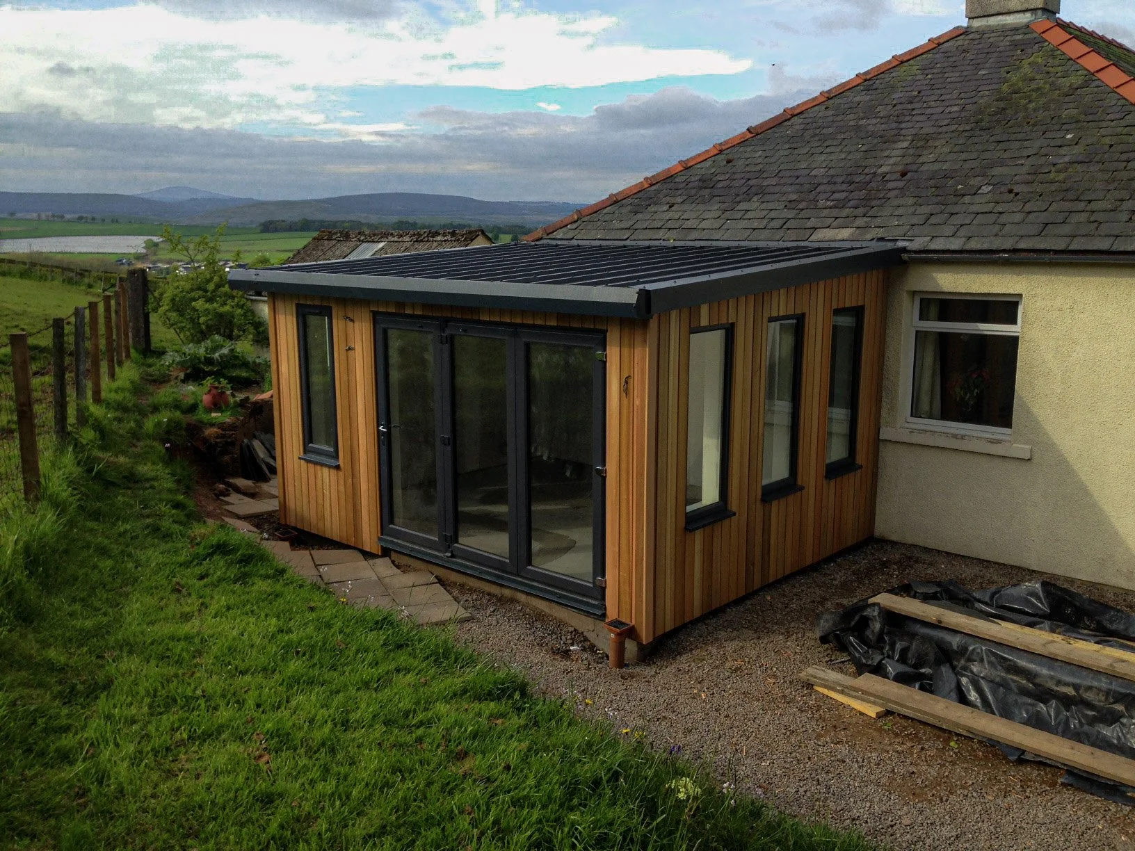 A small wooden extension with large glass doors and windows is built onto the side of a house with a shingled roof. The area around the extension is a mix of gravel and grass, with construction materials on the ground. In the background, there are hills and some open fields under a partly cloudy sky.