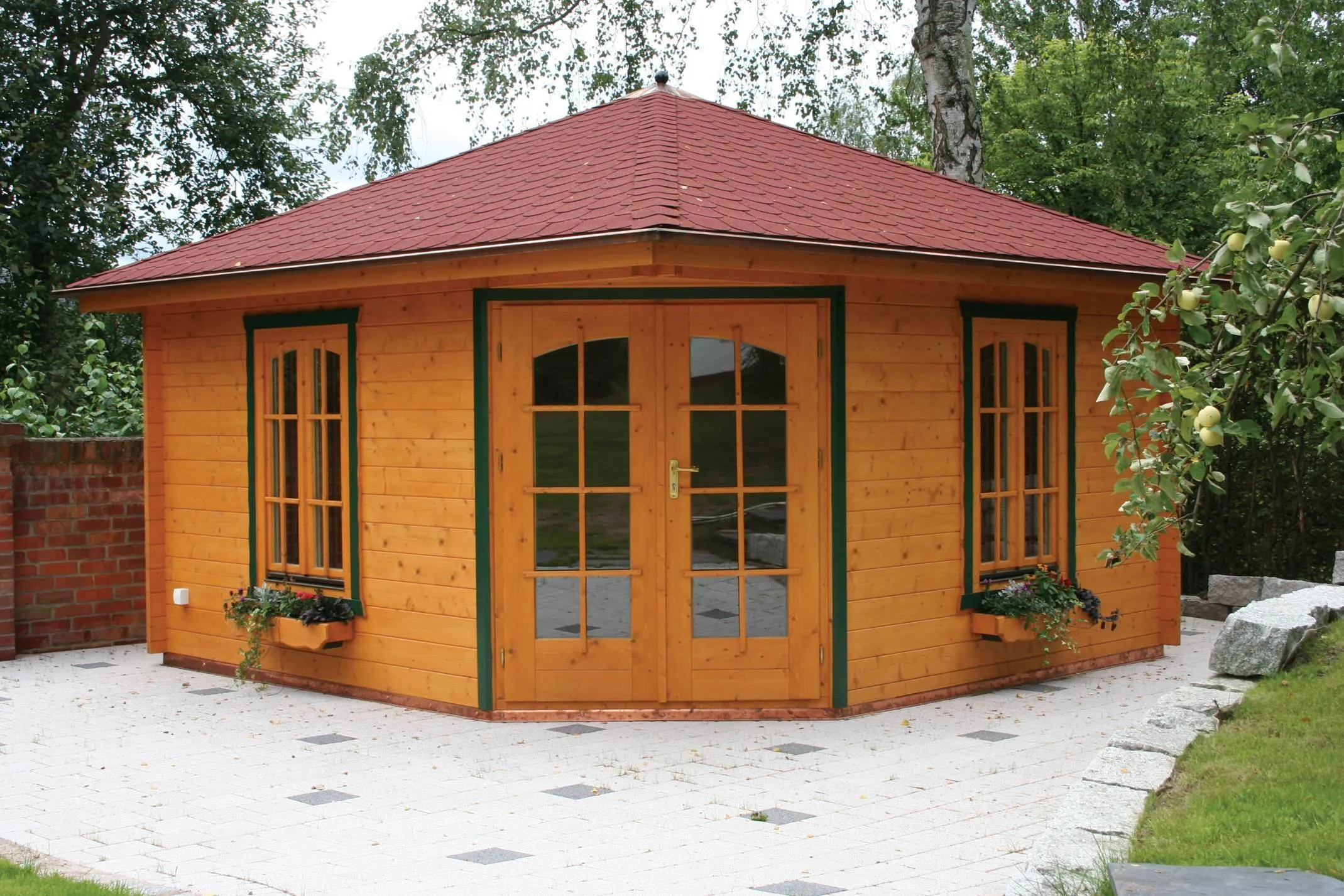 A small wooden garden shed with a red shingle roof, two windows with flower boxes, and double glass doors, situated on a white paved area surrounded by greenery.