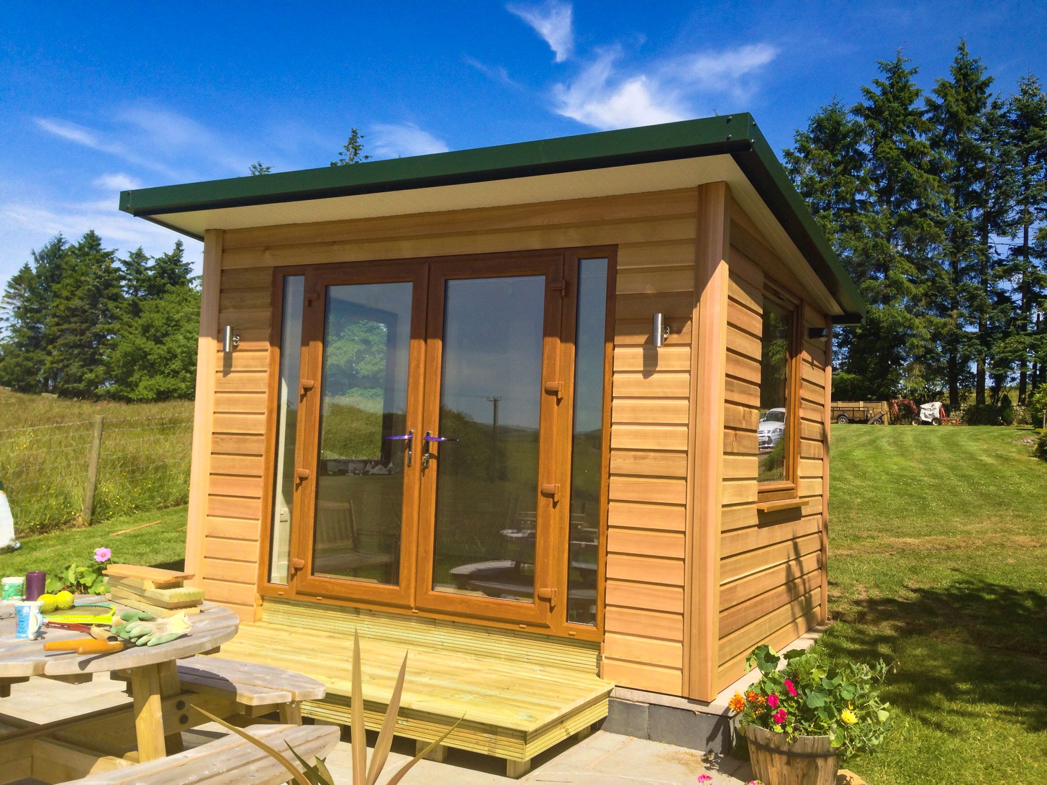 A small wooden garden shed with sliding glass doors and a window, set on a wooden deck, with gardening tools and colorful flowers nearby, under a bright blue sky.