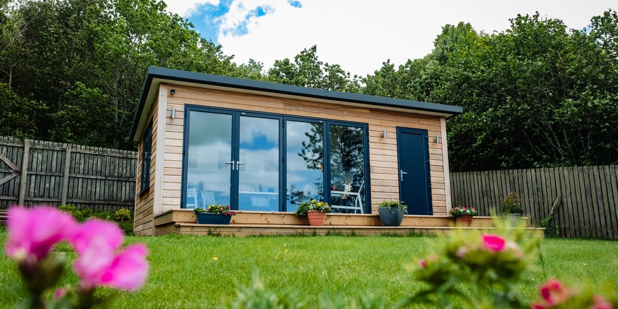 A small wooden garden house with blue framed glass doors and a blue door on a green lawn, surrounded by potted plants and flowers, with a wooden fence and trees in the background.