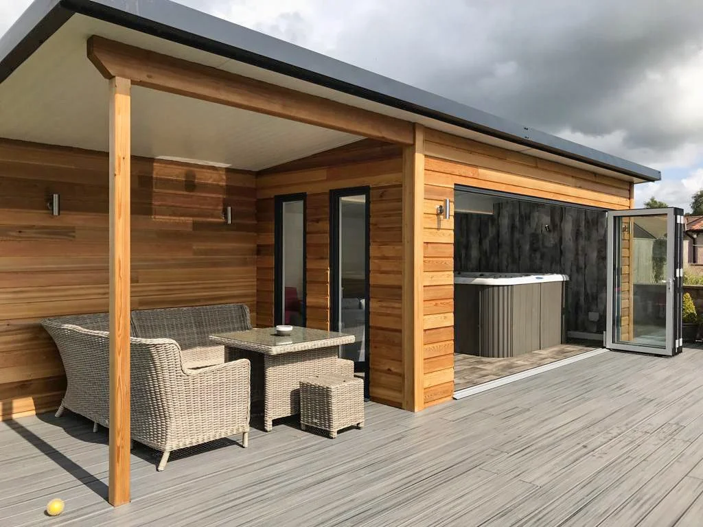Outdoor deck with patio furniture, including a glass-top table, wicker chairs, and an outdoor hot tub under a wooden overhang, with a partly cloudy sky above.