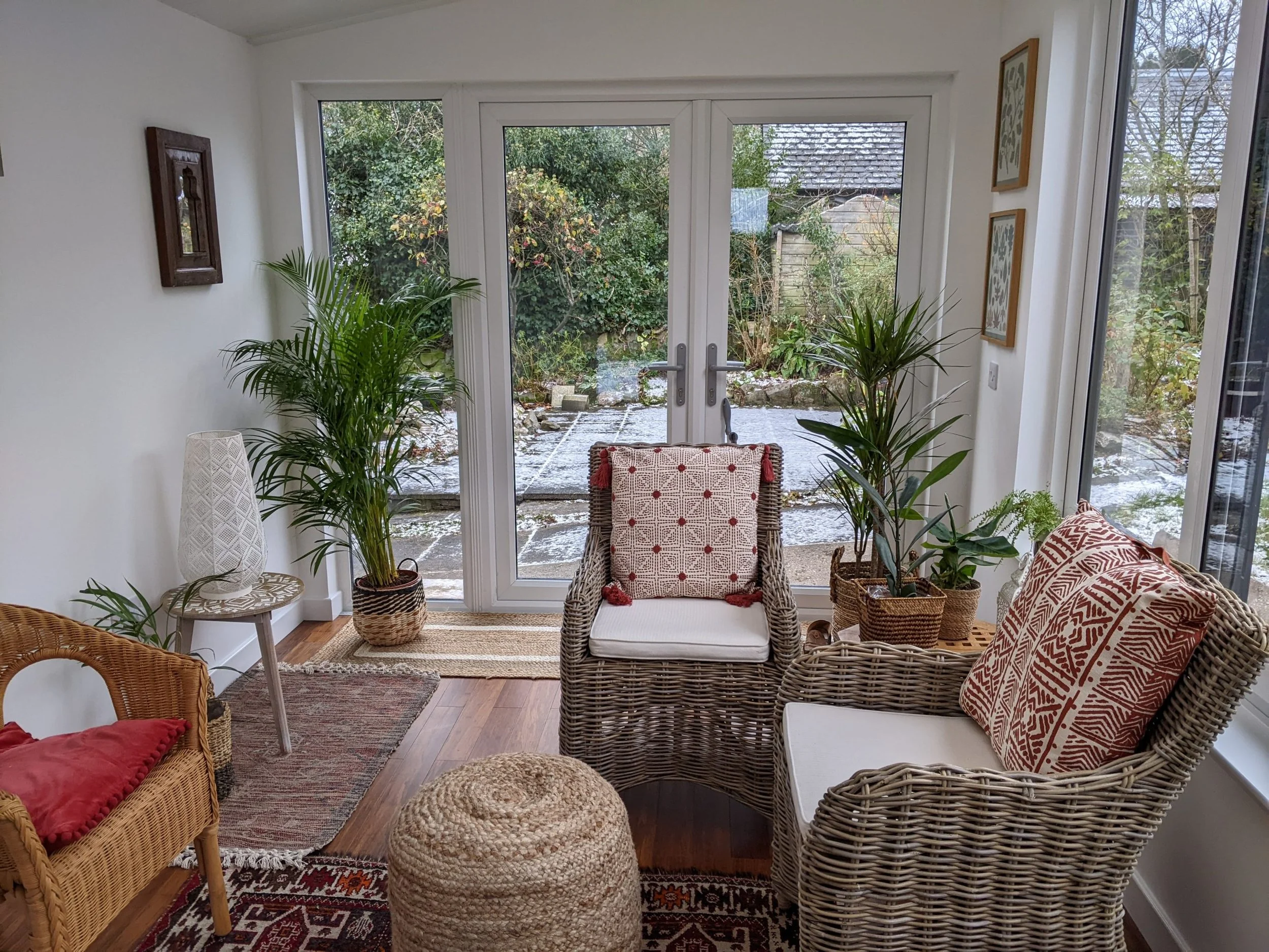 Sunroom with wicker and wooden chairs, decorative pillows, plants, and a view of a snowy garden outside.