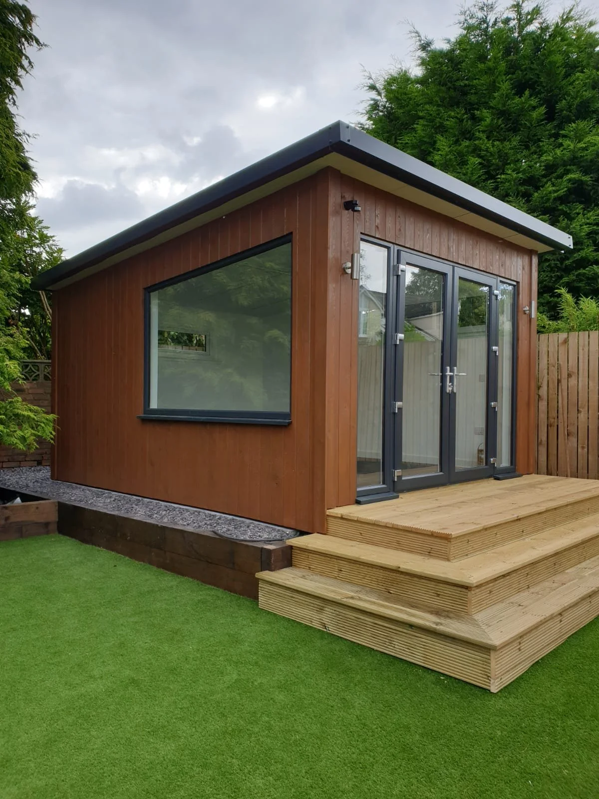 A small modern backyard shed with brown wooden siding, large glass double doors, and a big window, surrounded by green trees and a fenced yard, with a wooden deck and steps leading up to the entrance.