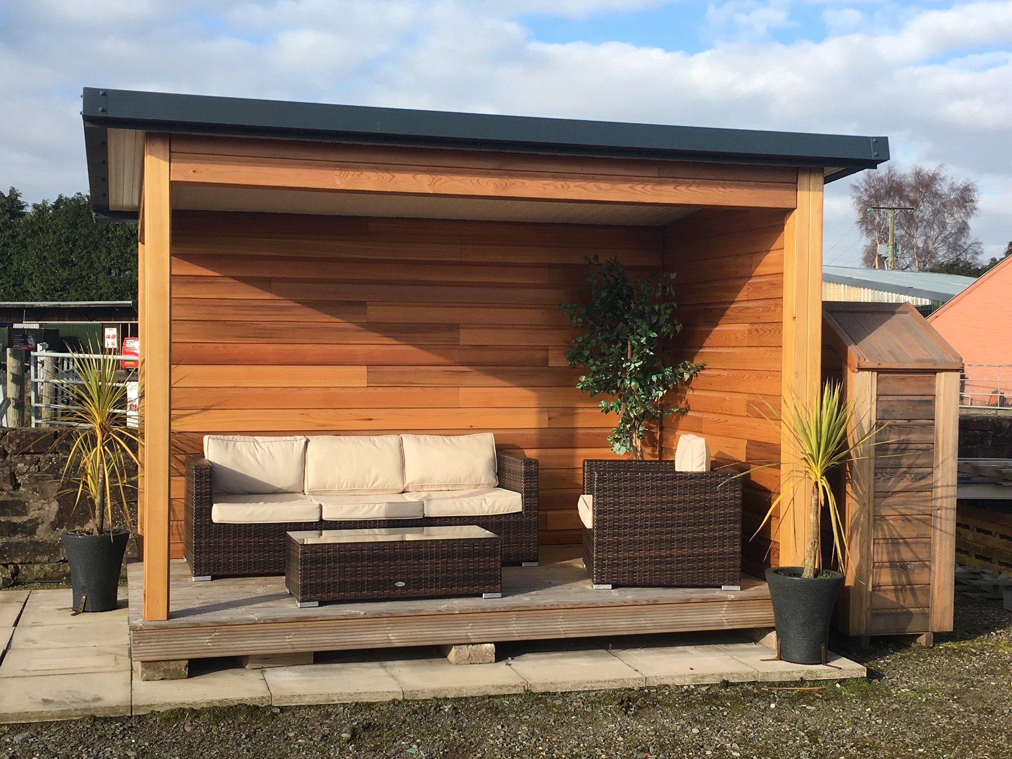 Outdoor wooden patio with beige cushioned sofa and armchair, small glass-topped table, potted plants, and a wooden privacy wall, under cloudy sky.