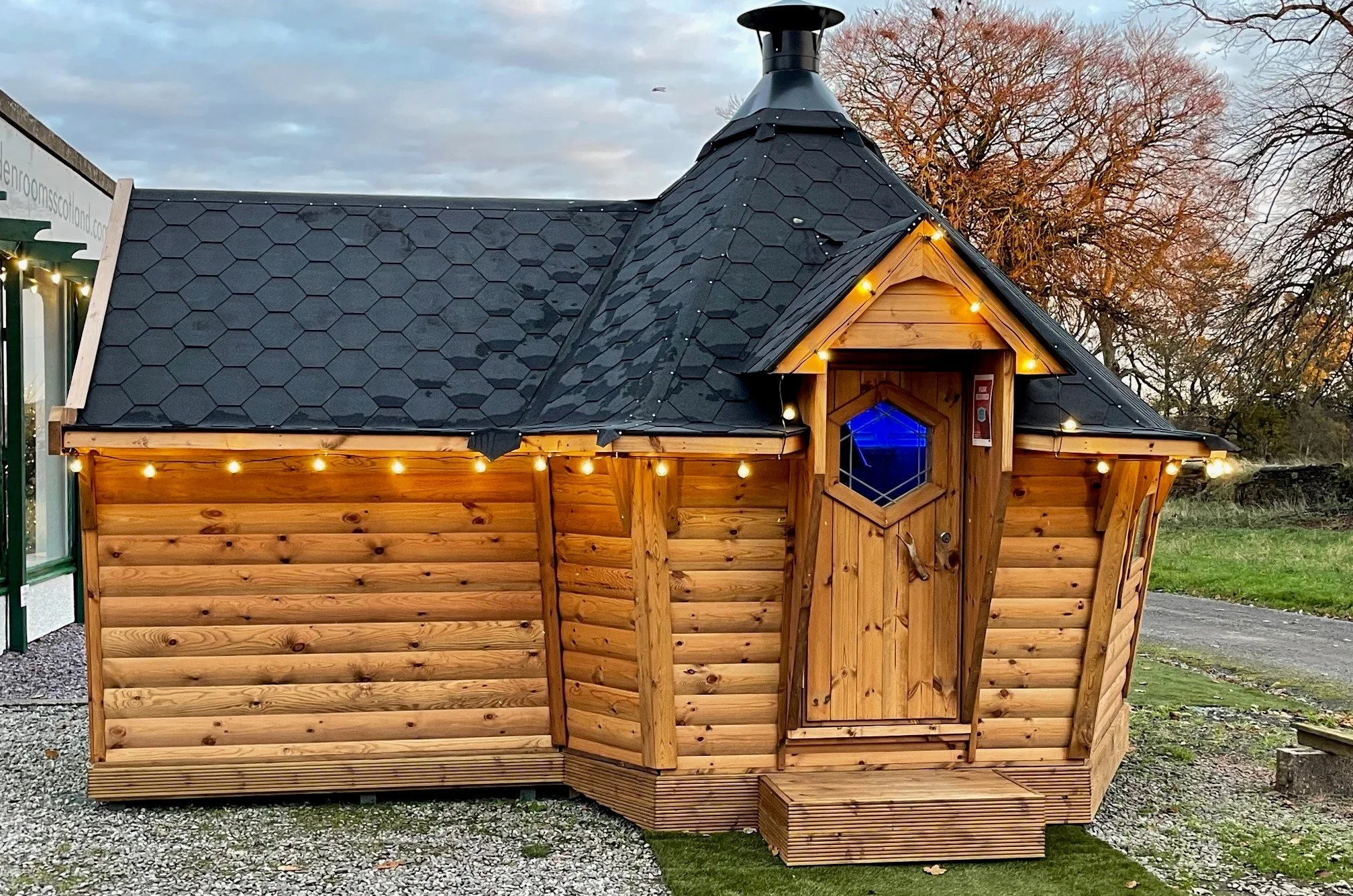 A small wooden cabin with a hexagonal window, black shingled roof, and string lights along the roof edges. There is a wooden door with a small step leading up to it. In the background, there are bare trees and a grassy area.