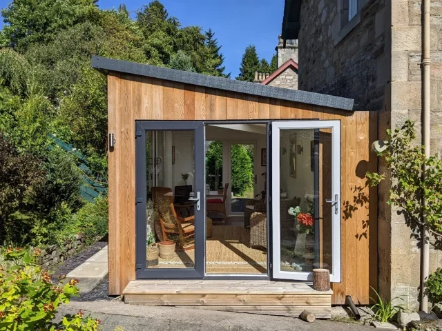 Small wooden sunroom with large glass doors, attached to a house, surrounded by greenery.