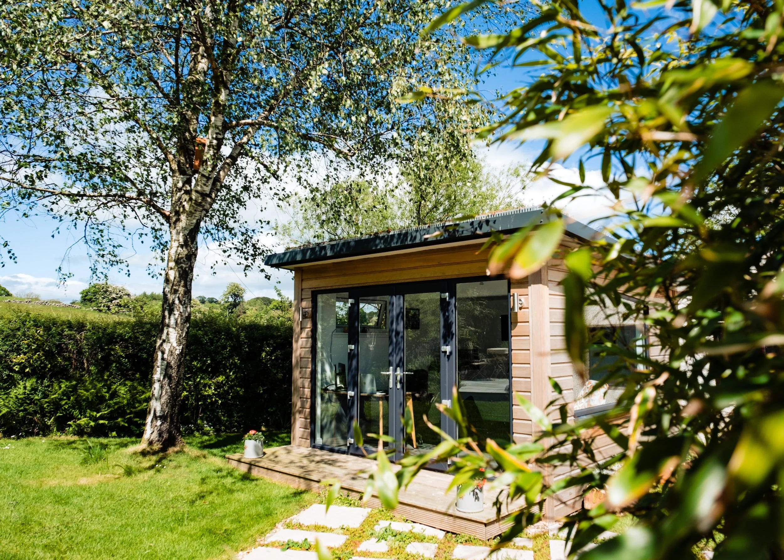 A small wooden garden shed with glass double doors, situated on a grassy lawn with trees and bushes around it, under a blue sky.