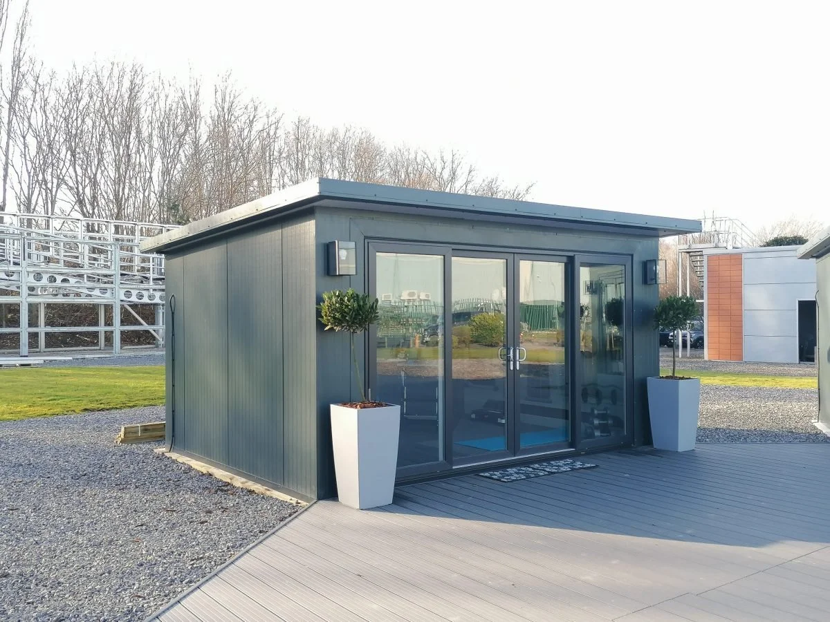 Small modern gray building with large glass sliding doors, two tall white planters with green shrubs, and an outdoor deck area.
