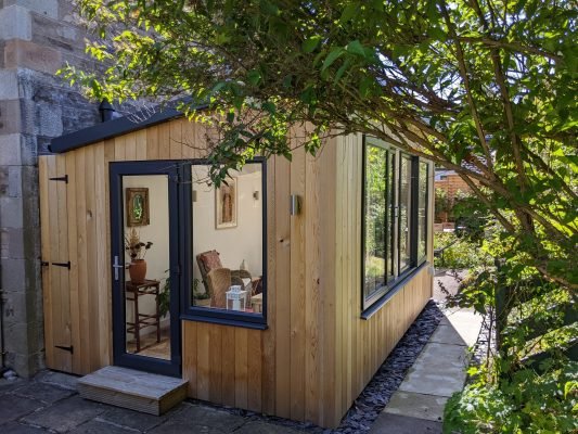 Tiny wooden house with large windows and a door, surrounded by greenery and trees.