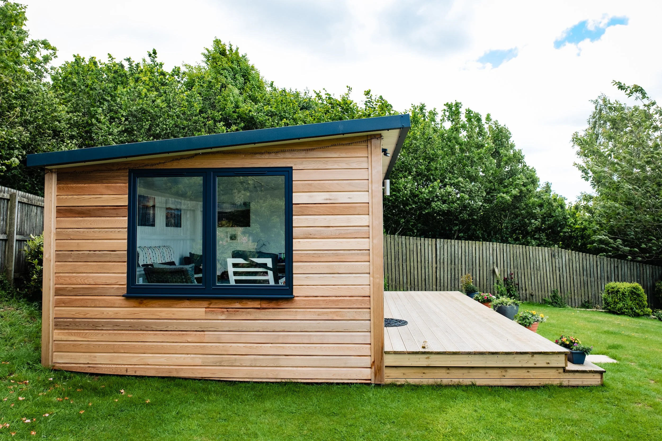 A small wooden garden shed with a large window, located in a green backyard with potted plants on a wooden deck and a wooden fence in the background.