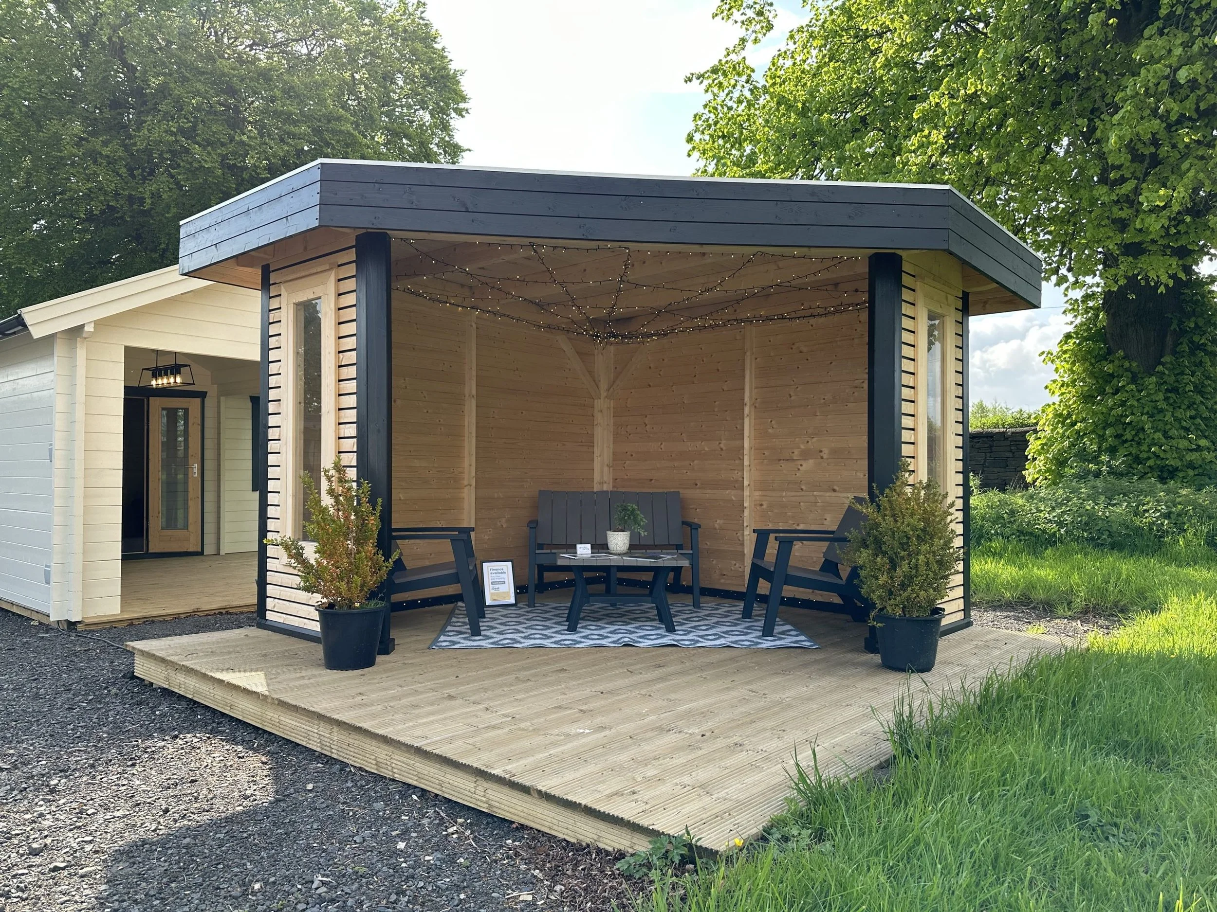 An outdoor wooden pavilion with string lights, chairs, a small table, potted plants, over a wooden deck, with a neighboring house in the background and green trees.