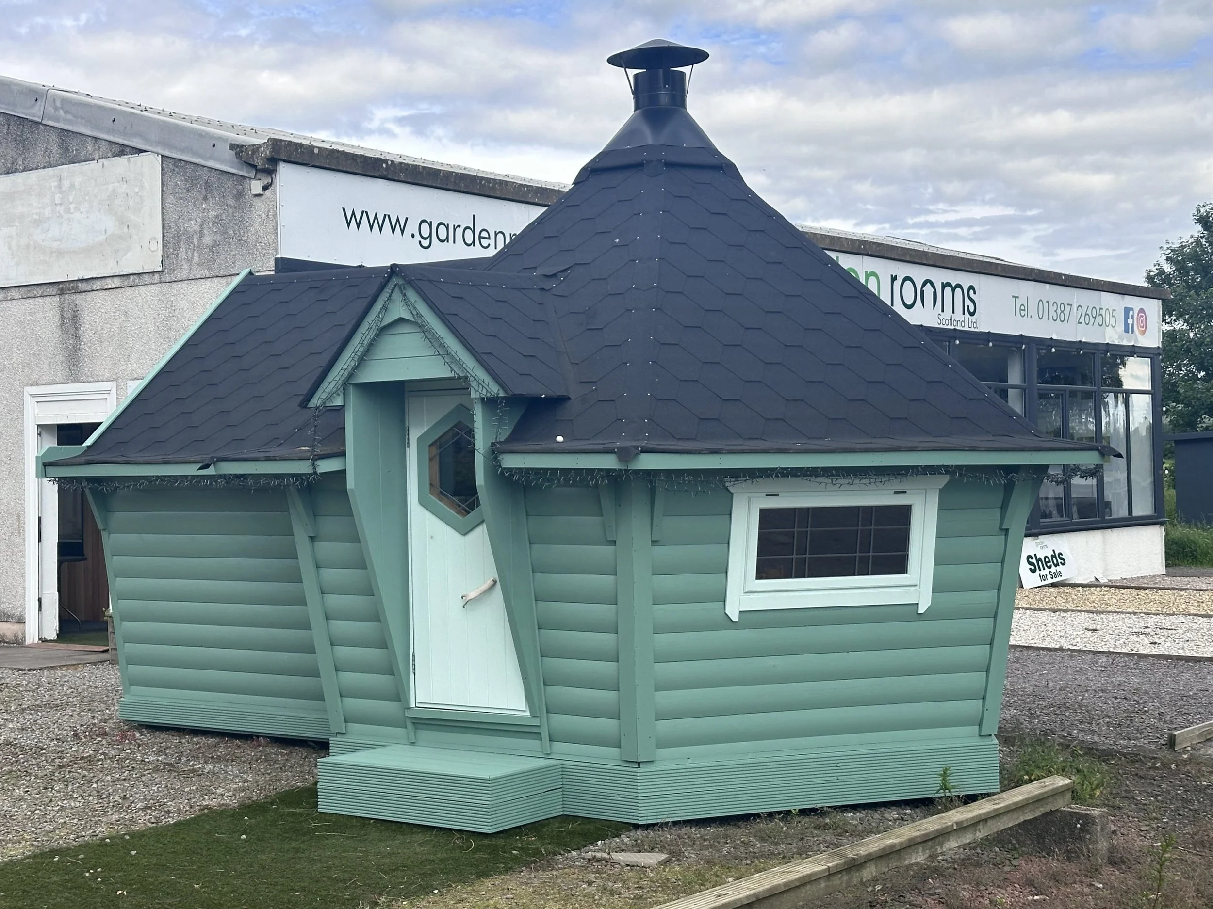 A small, mint green wooden cabin with a unique tilted and rounded shape, black shingled cone roof, window, and white door, situated outdoors on a gravel lot.