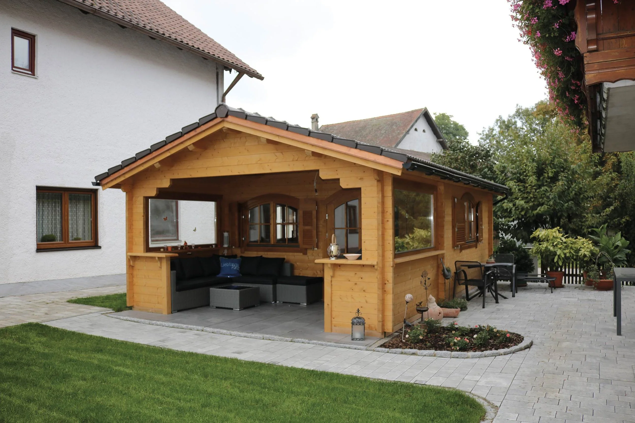 Wooden garden pavilion with seating area, surrounded by a paved patio and a small garden bed with flowers, adjacent to a white house with brown-framed windows.