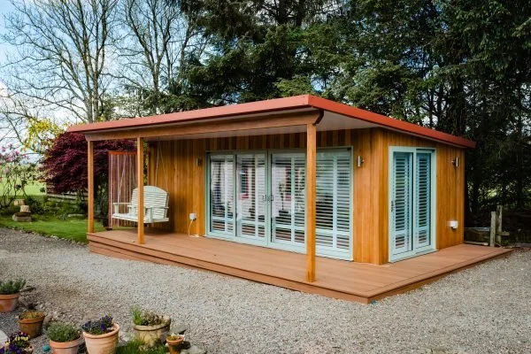 A small wooden house with large glass sliding doors, a covered porch with a white swing, situated on a gravel lot surrounded by trees and potted plants.
