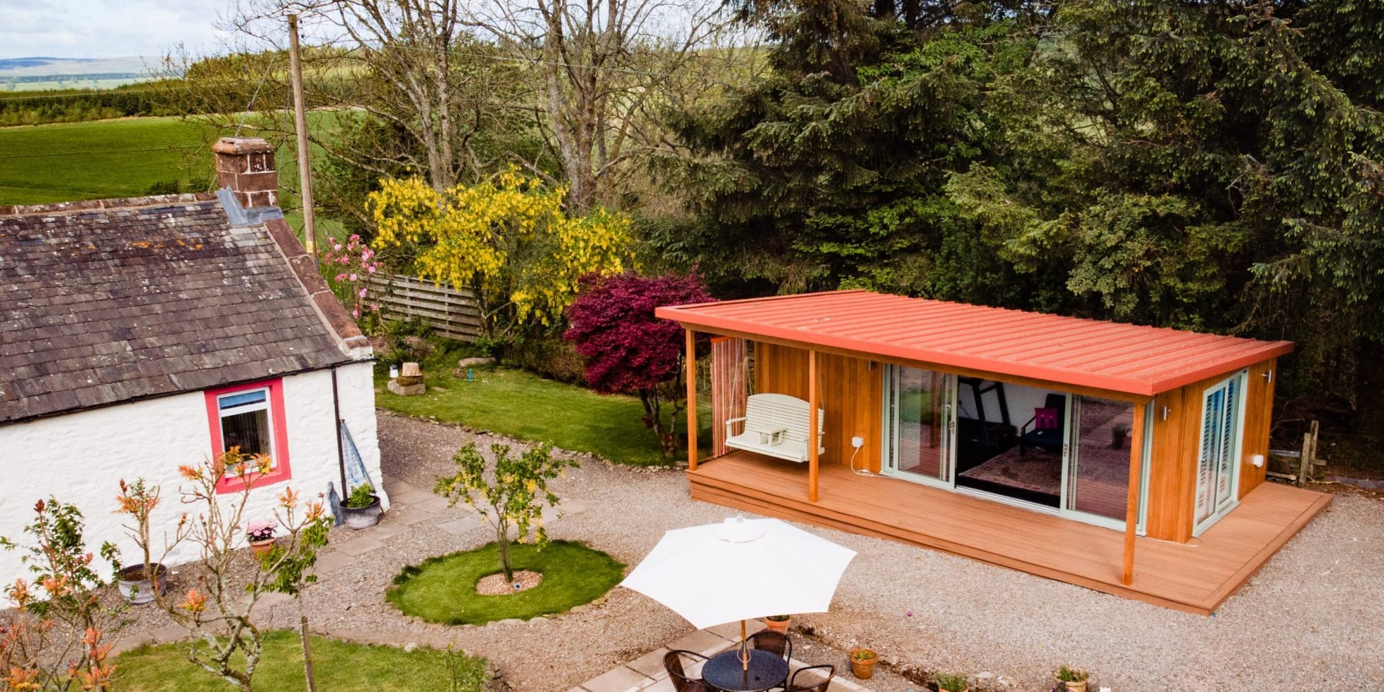 A backyard scene with a white cottage that has pink window trim, a garden with trees and plants, and a modern wooden extension with sliding glass doors, a porch swing, and a red metal roof. There is an outdoor table with an umbrella and chairs, surrounded by a gravel yard and lush green trees.