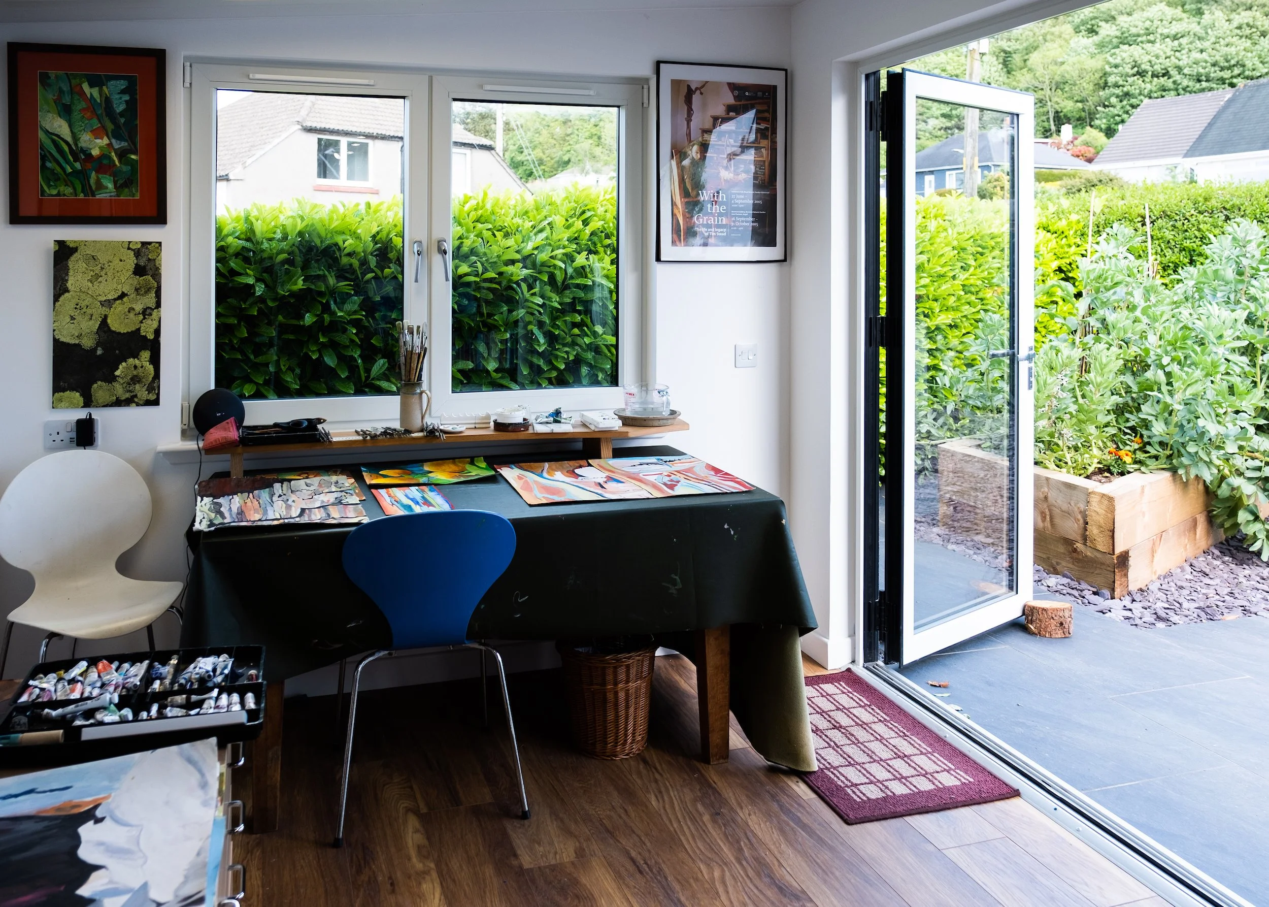 An art studio with a black table covered in colorful paintings, a white and a blue chair, a window and a glass-door leading to a garden with green bushes and raised plant beds.
