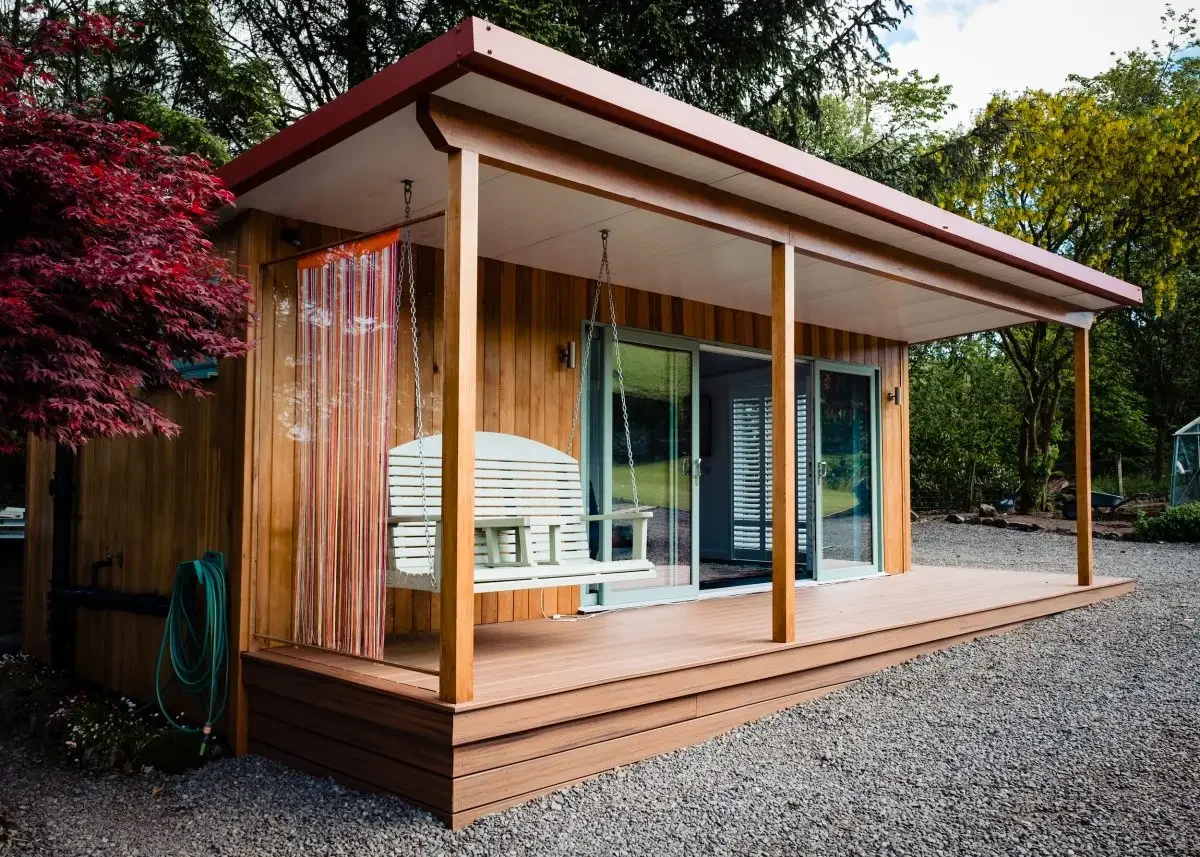 Small wooden house with a covered porch, white swing, sliding glass doors, and a gravel yard, surrounded by trees.