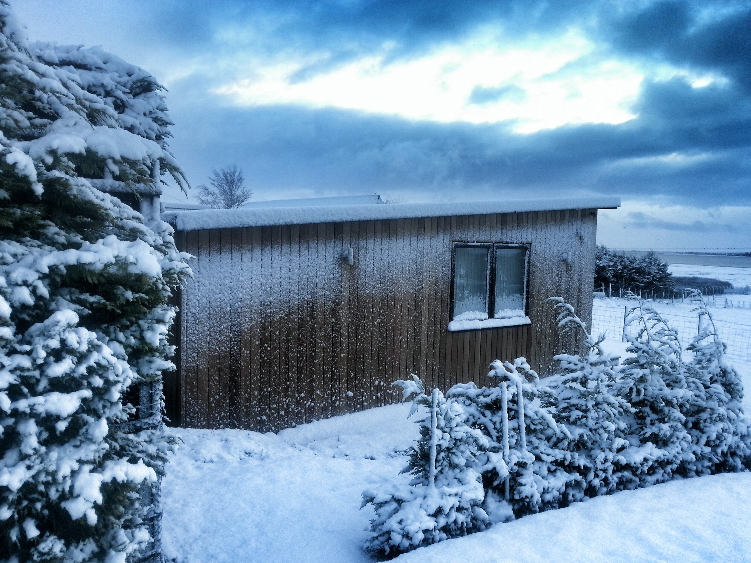 Snow-covered house with wooden exterior, windows with snow on the sills, surrounded by snow-laden bushes and trees. The sky is cloudy with patches of light.