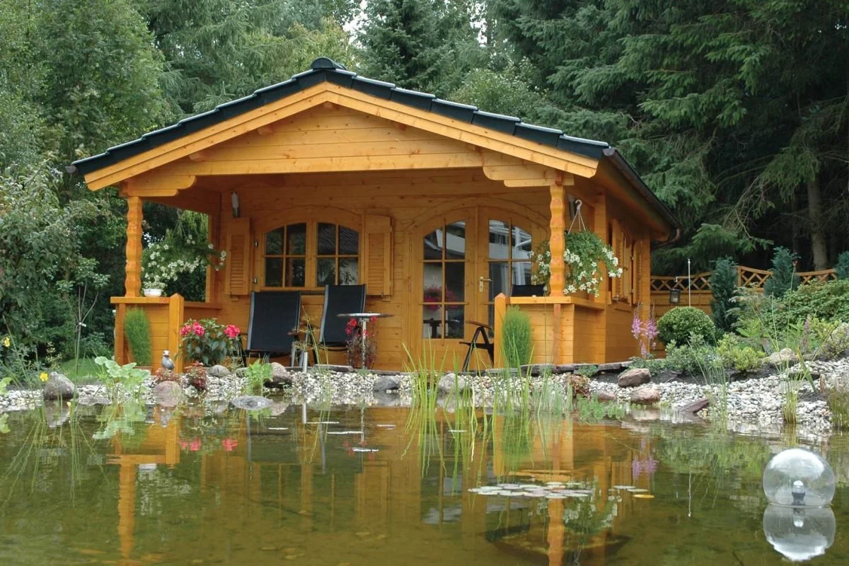 A wooden cabin with a black roof, surrounded by trees, facing a pond with water lilies and rocks, with chairs and hanging plants on the porch.
