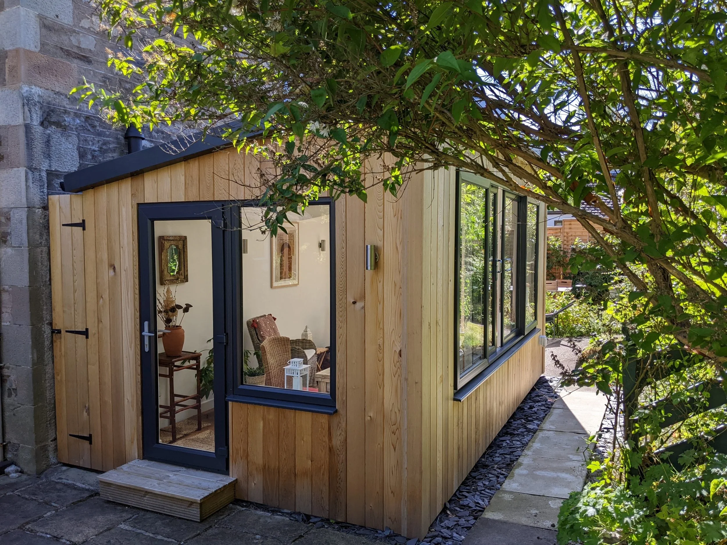 Small wooden backyard shed with large windows, black framed door and door step, surrounded by green foliage.