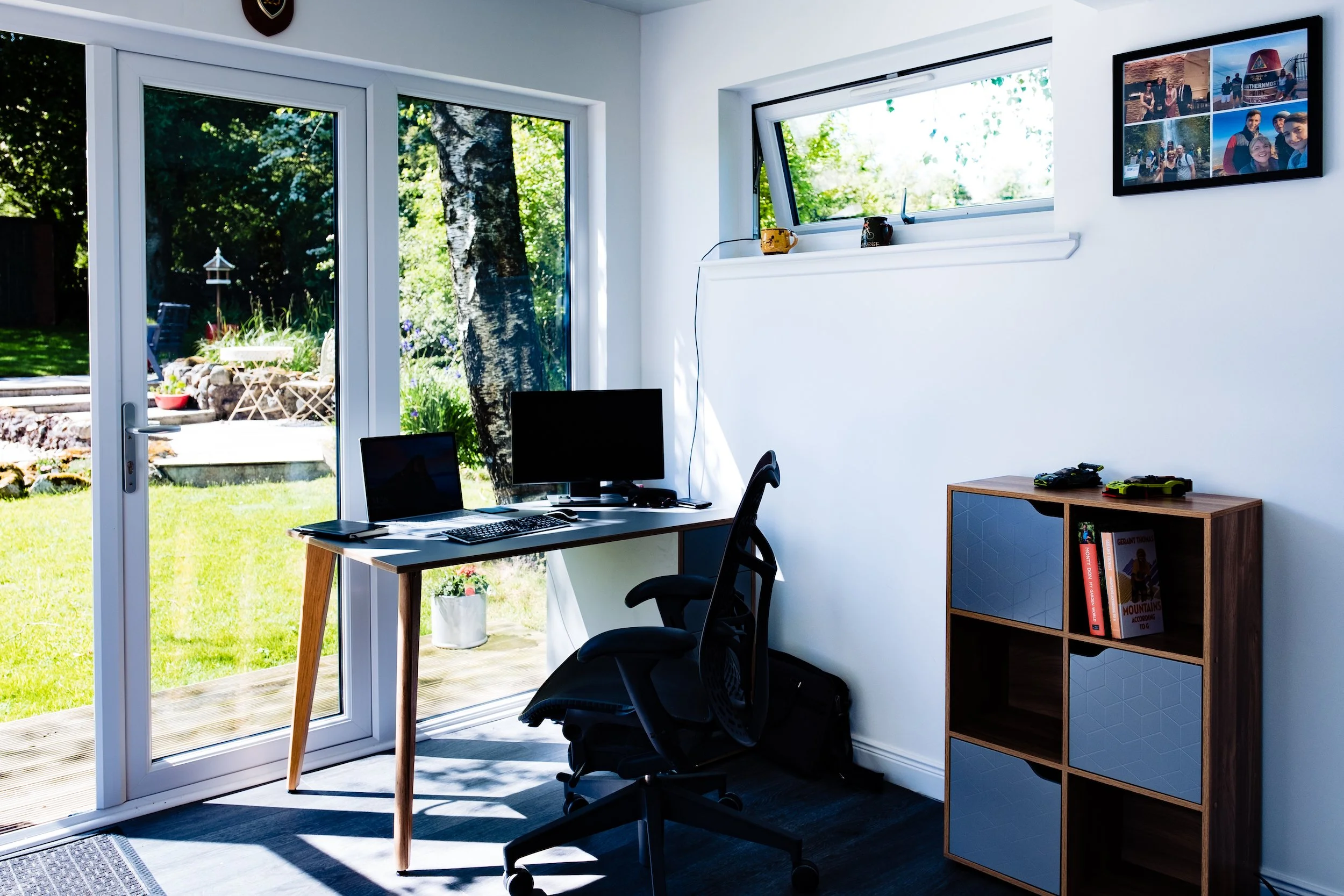 Home office with desk, chair, two monitors, laptop, and a sliding glass door leading to a backyard garden.